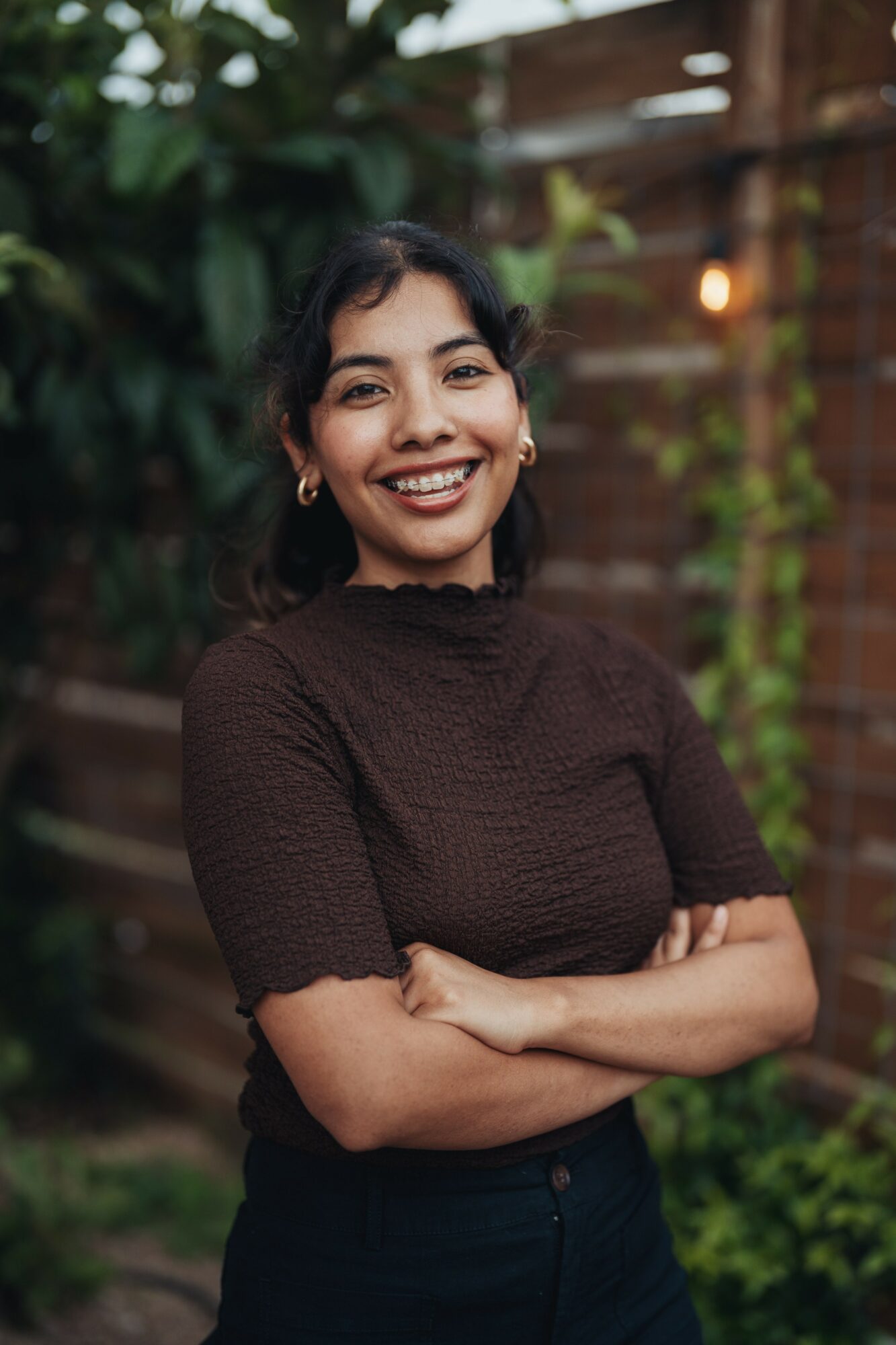 Young woman with dark hair smiling, arms crossed, outdoors with greenery and wooden fence in background.