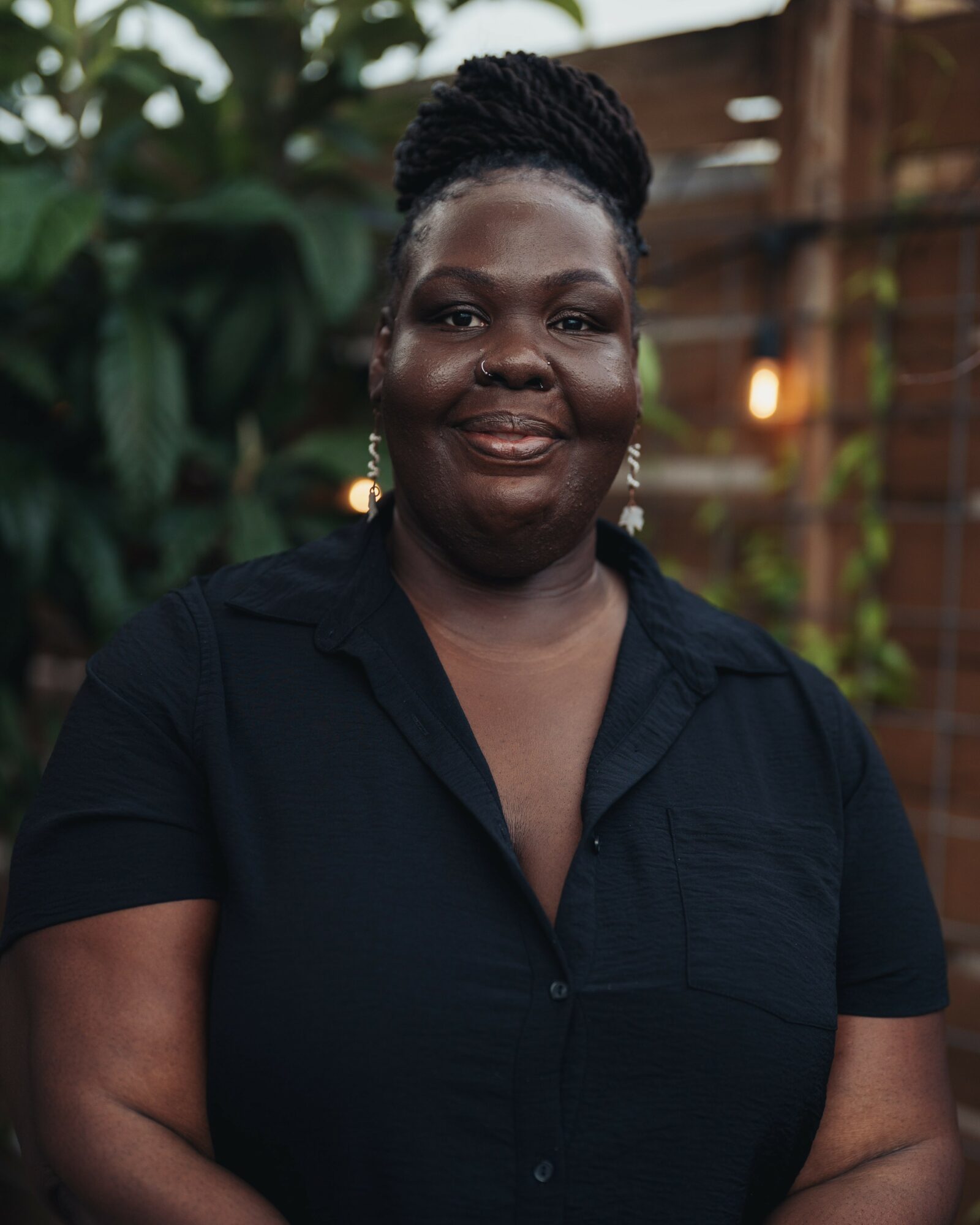 Portrait of a smiling woman with braided hair, wearing a black shirt, outdoors with greenery and wooden background