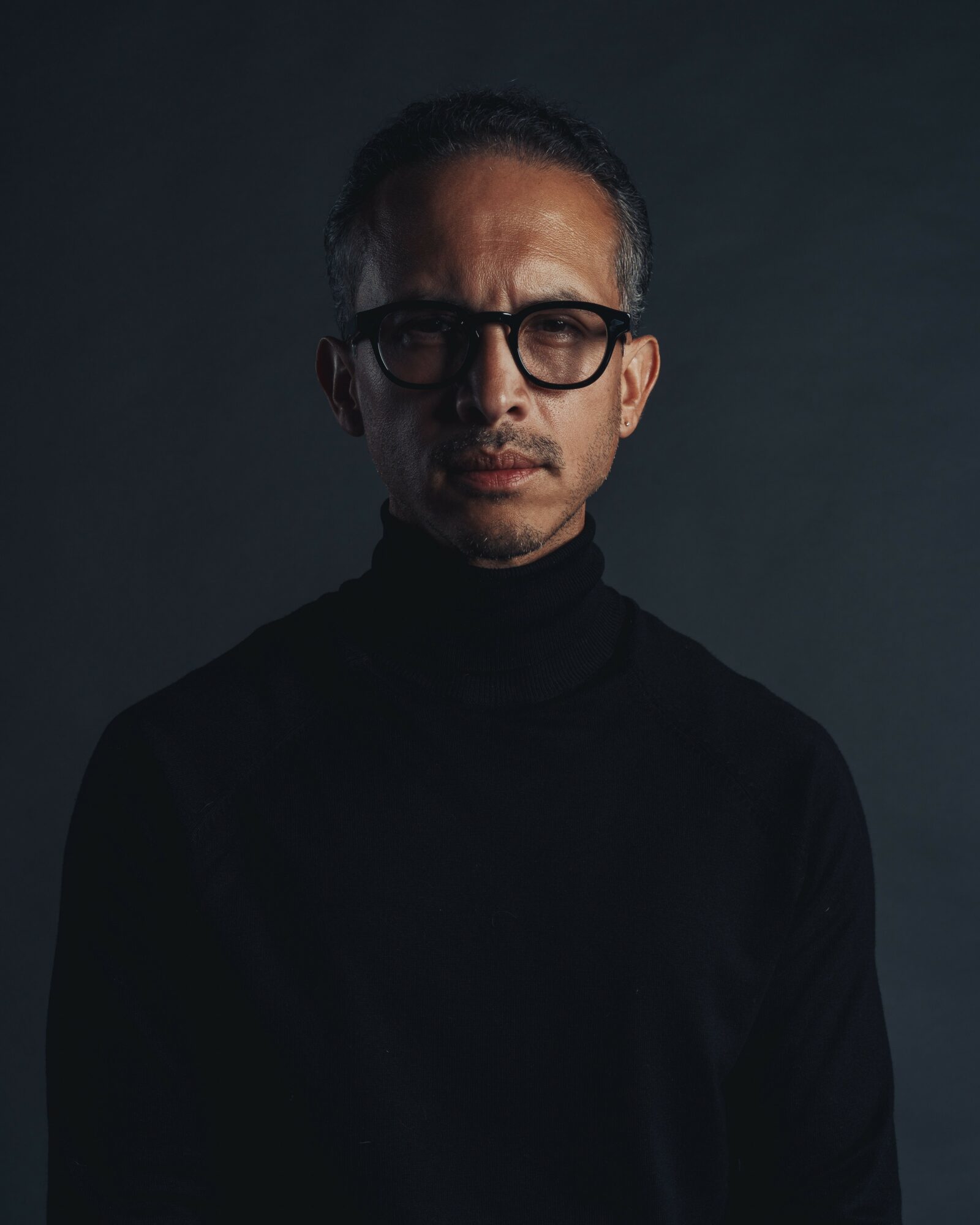 Man with glasses and short hair wearing a black turtleneck, looking at the camera against a dark background.