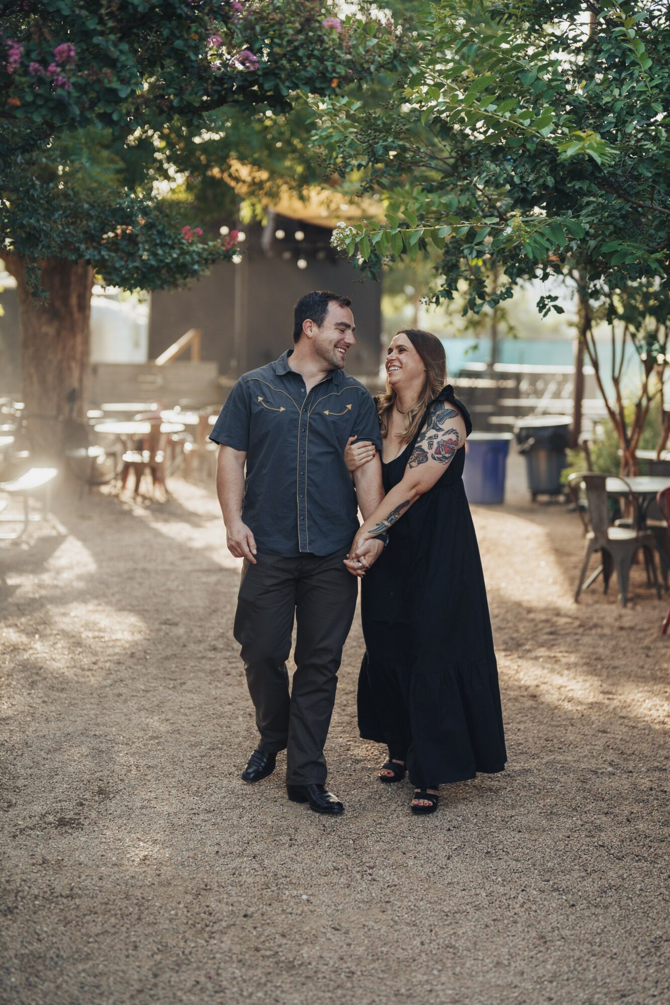 A man and woman walk hand in hand outdoors under trees, smiling at each other, with tables and chairs in the background.
