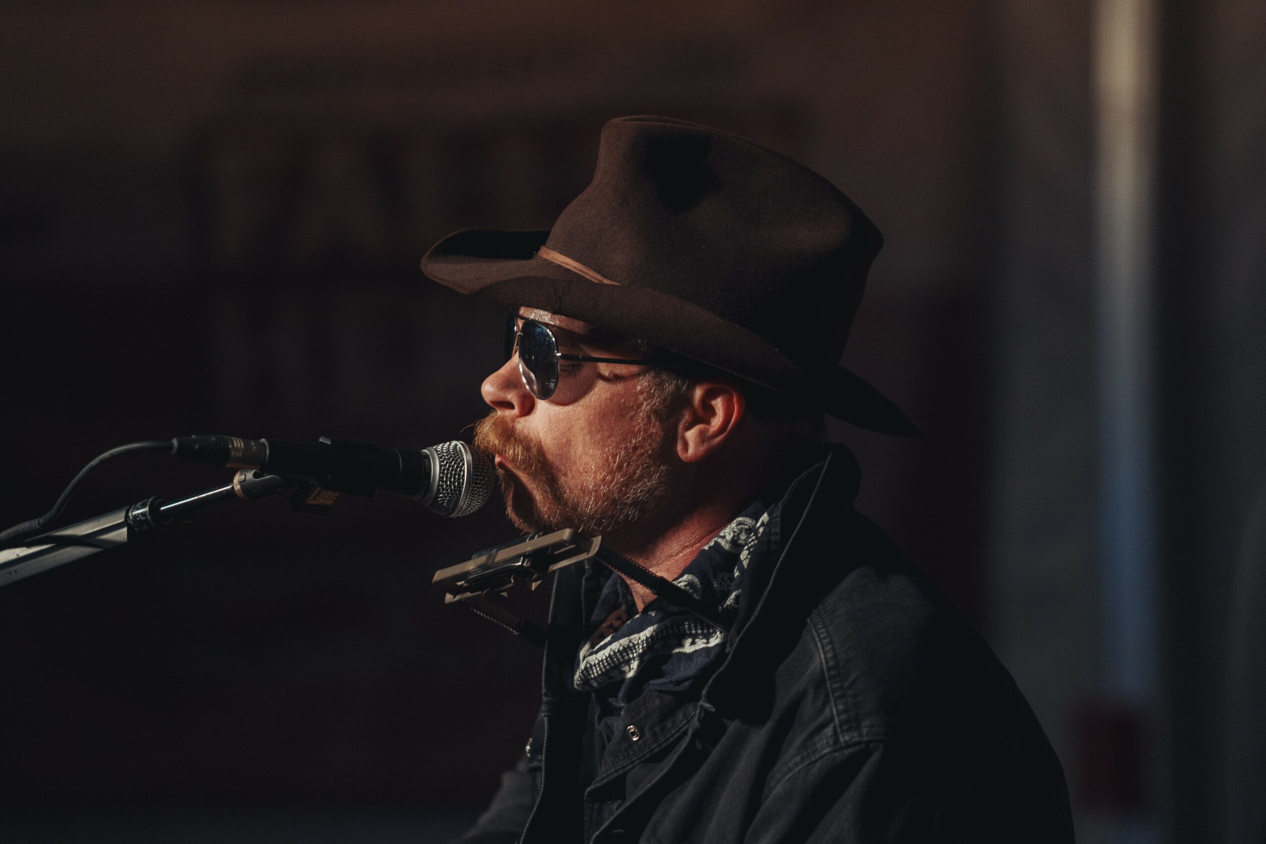 Man wearing a hat and sunglasses singing into a microphone, with a dark background.