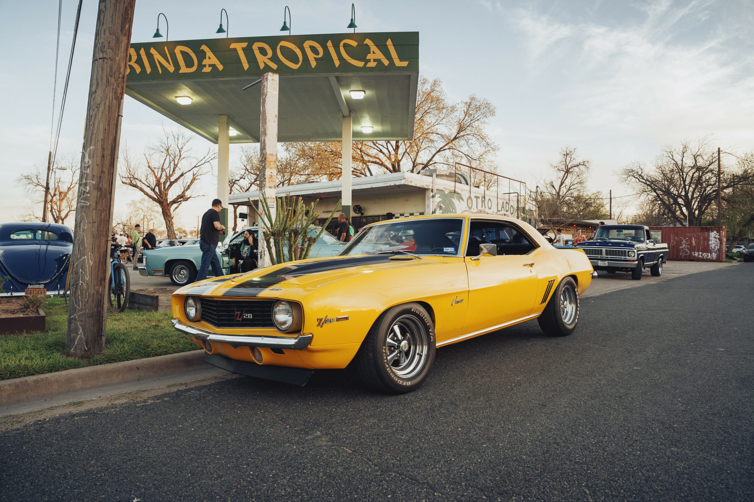 Yellow vintage muscle car parked on street near a tropical-themed sign and other vehicles, trees, and sky in background.
