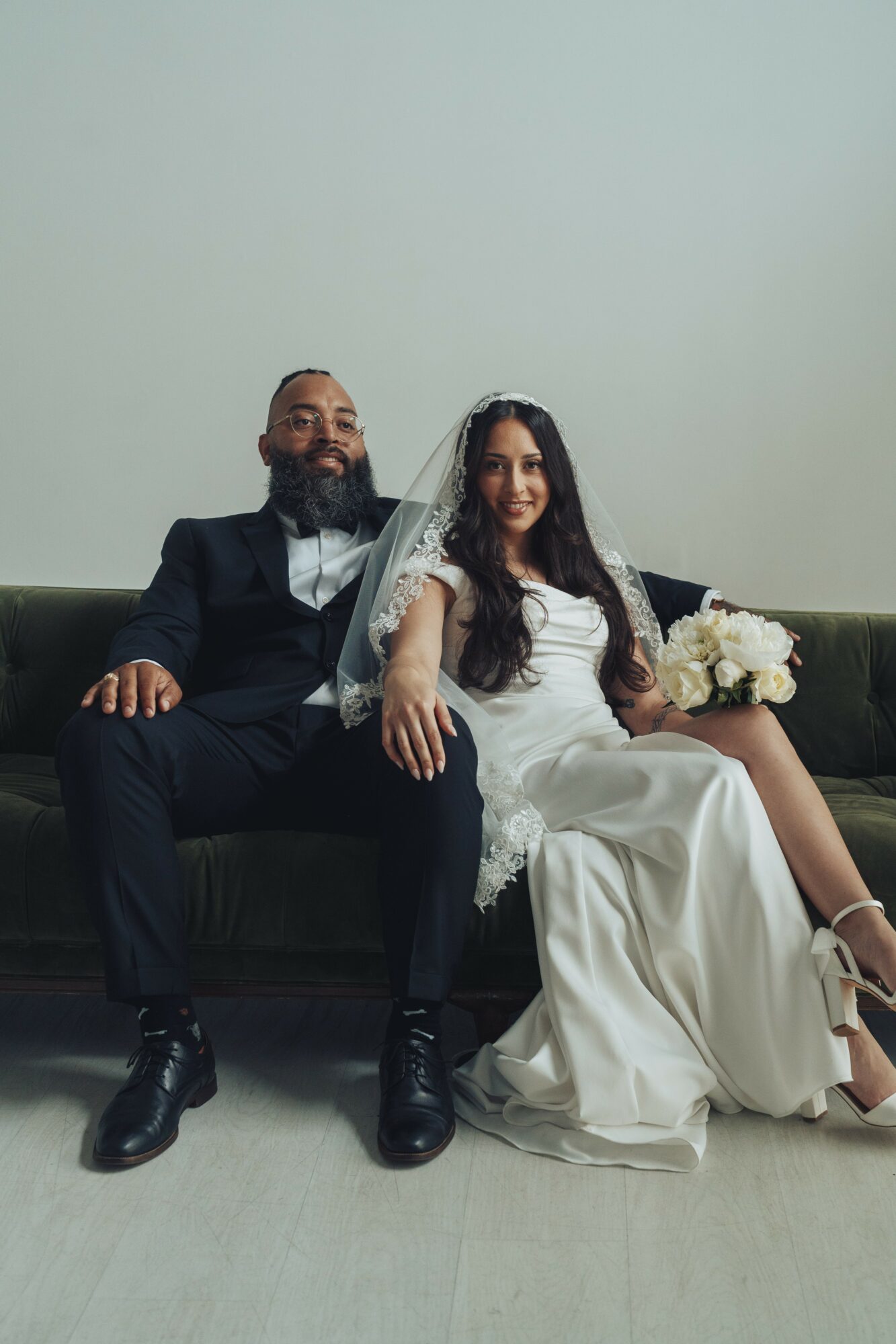 A bride and groom sit on a dark green sofa, smiling, with the bride holding a bouquet of flowers.