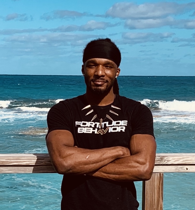 Man with arms crossed standing on a beach with ocean and sky in background.