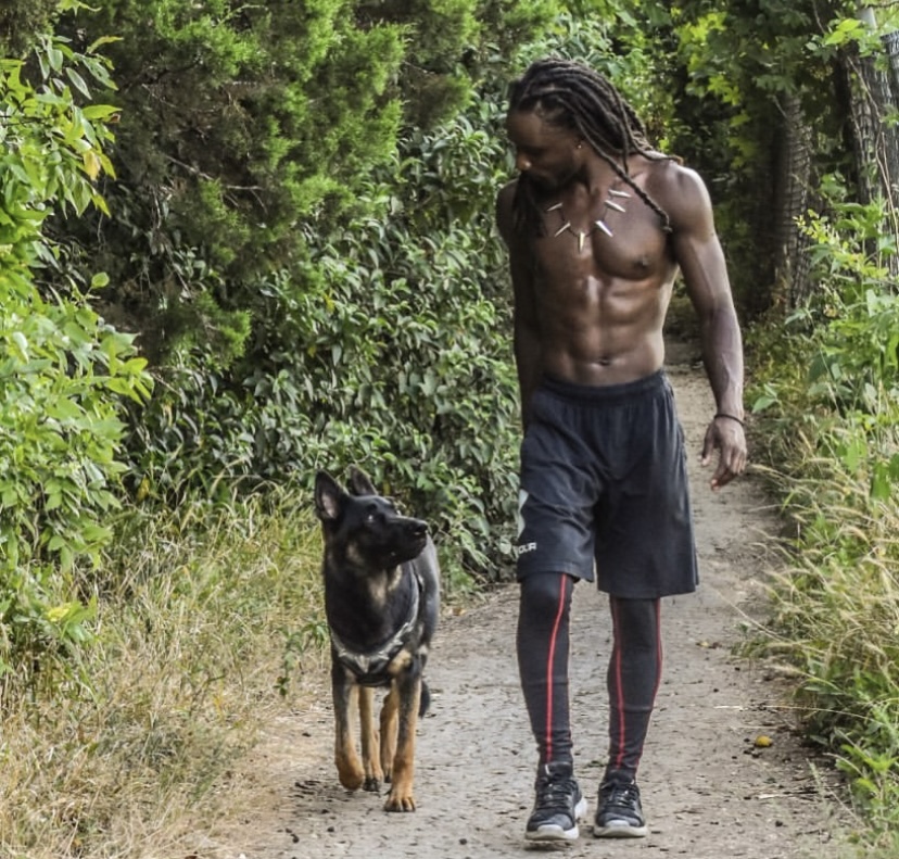 A shirtless man with dreadlocks walks on a dirt path with a black and tan dog in a green forest.