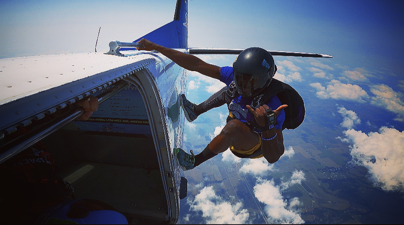 Person exiting an aircraft mid-air, sky and clouds in background, wearing helmet and safety gear, holding a peace sign.