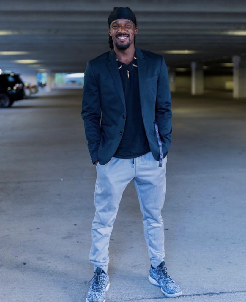 Smiling man in a dark jacket, black shirt, light pants, and sneakers standing in a parking garage.