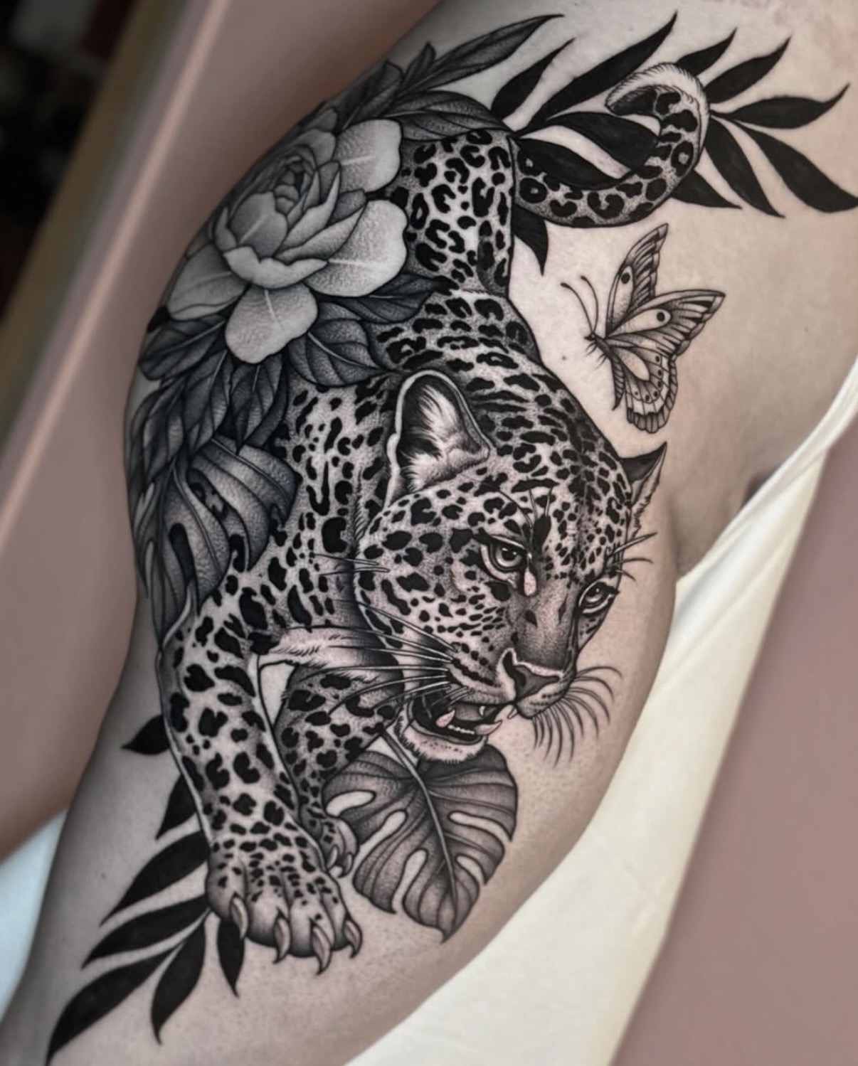 Leopard cub with flowers, leaves, and a butterfly, in black and gray tattoo style on skin.