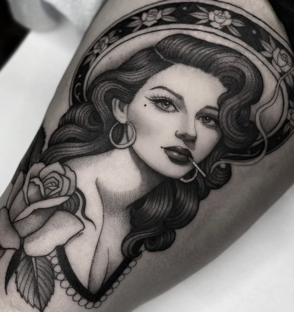 Black and gray tattoo of a woman with wavy hair, hoop earrings, a rose, and floral details on her hat.