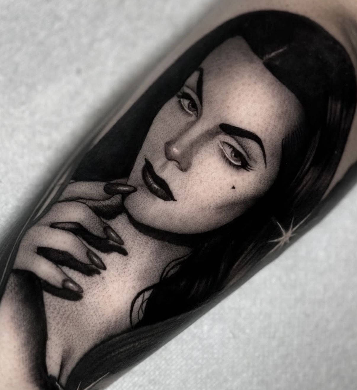 Black and white portrait of a woman with dark hair, makeup, and a confident expression, touching her face with her hand.