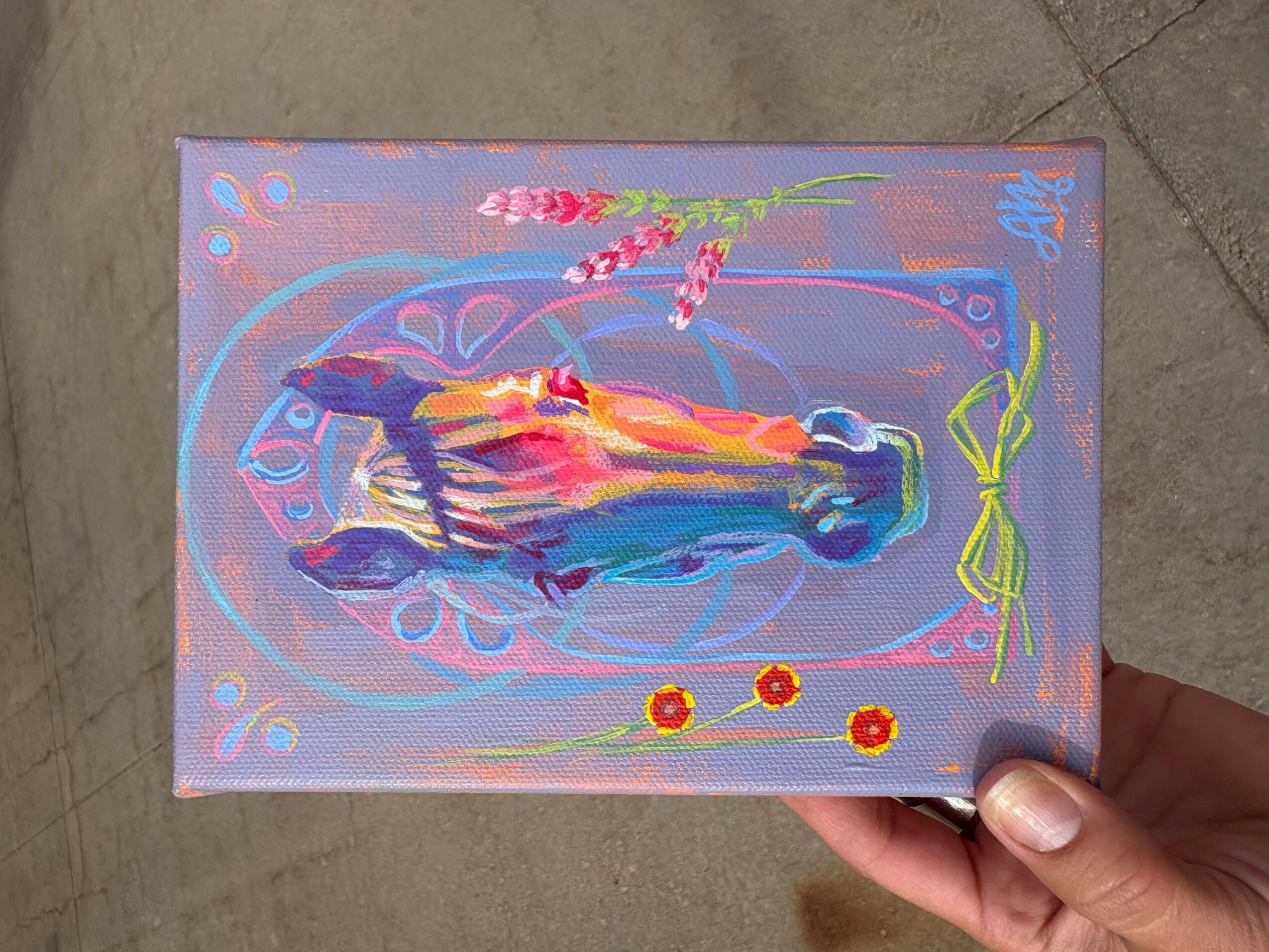 Hand holding a colorful painting of a fish with flowers and decorative elements on a purple background.