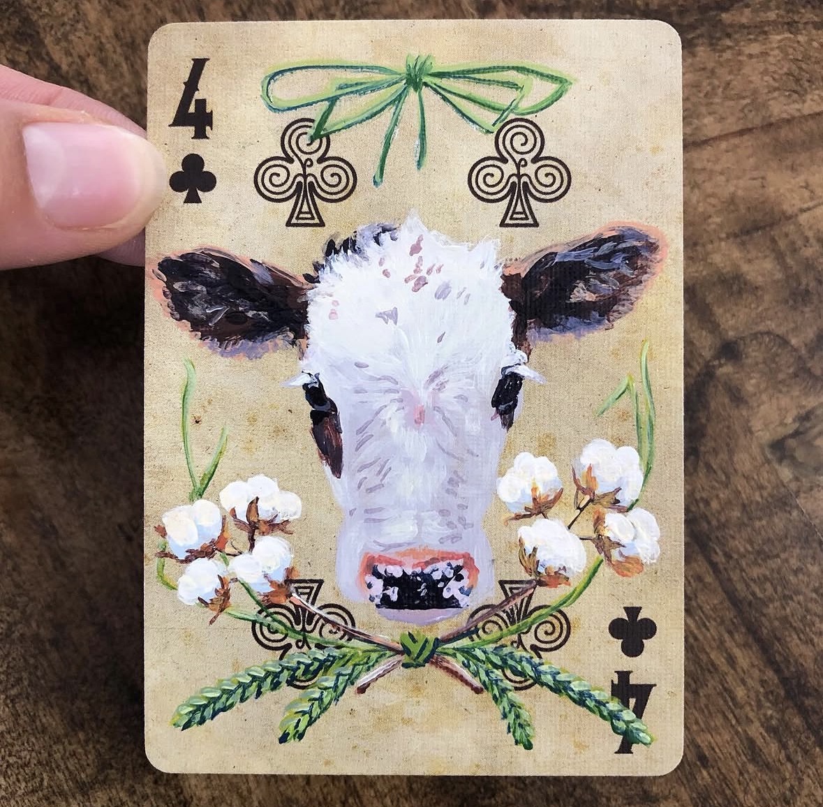 Playing card four of clubs with a cow's face and floral decorations, including cotton flowers and green leaves.