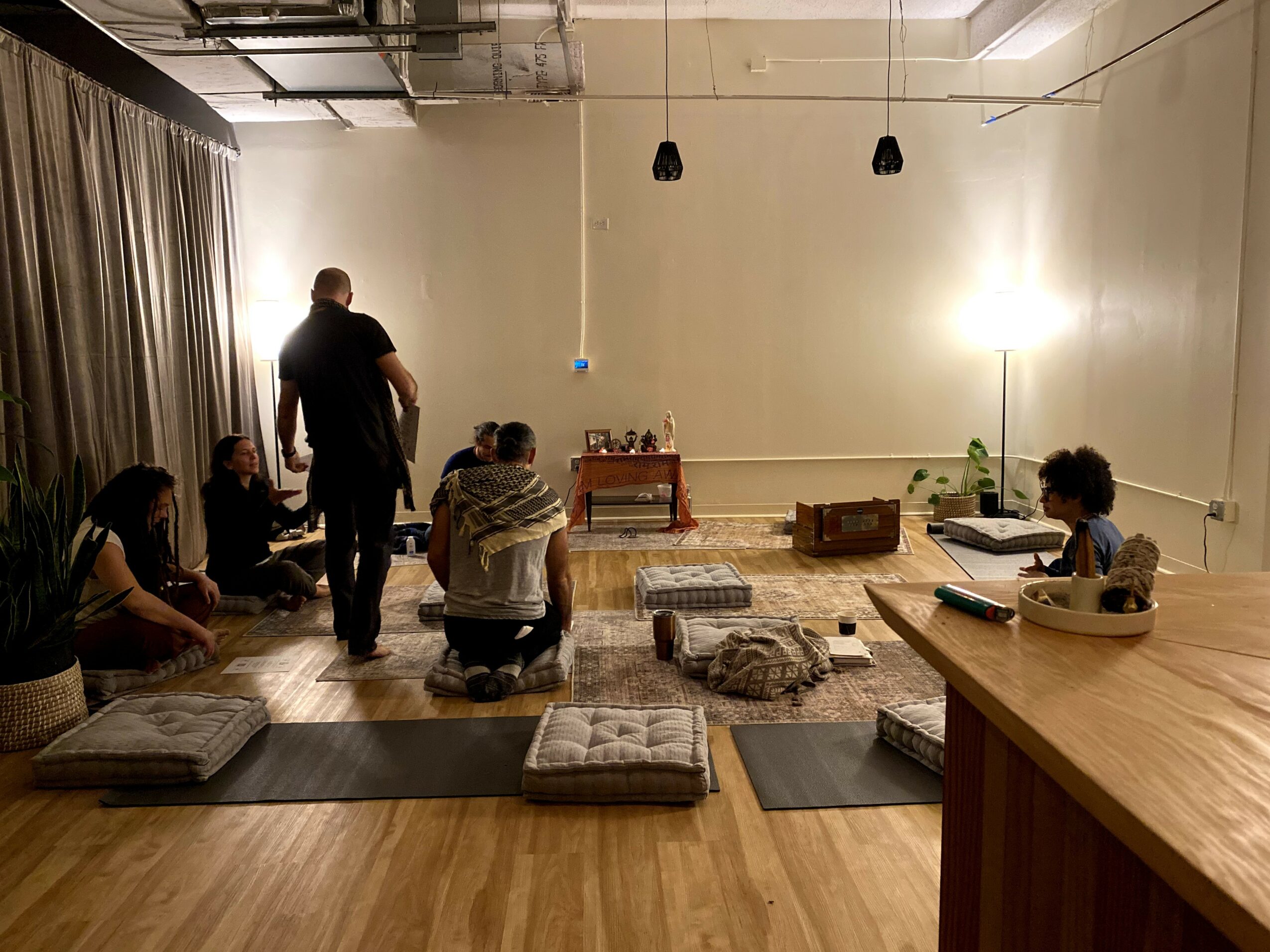 People sitting on cushions in a room with a wooden floor, some standing, with a table and a lamp in the background.