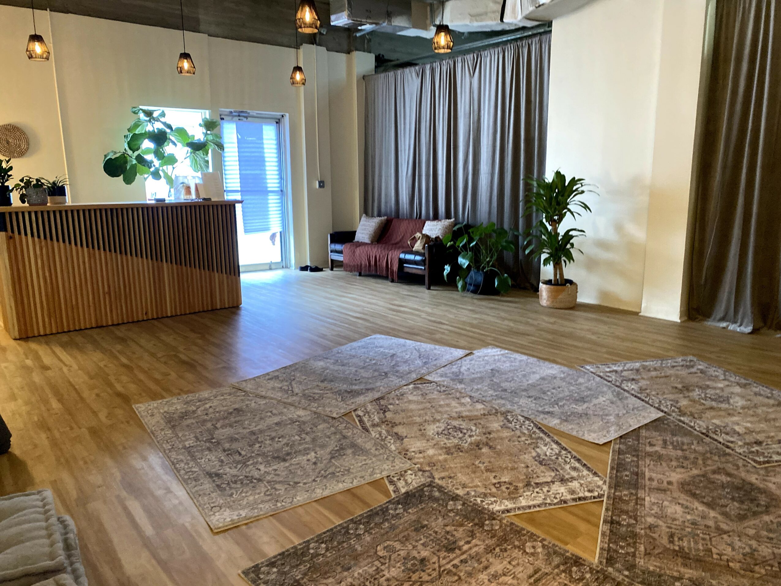 Interior space with wooden flooring, a curved reception desk, seating area with plants, and large curtains, well-lit with hanging lights.