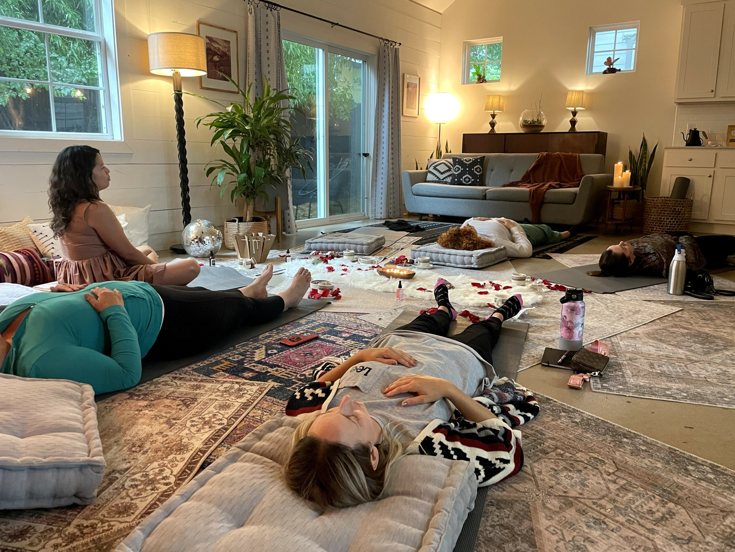 People relaxing in a living room with rugs, furniture, and plants, some lying down and others sitting, with natural light coming through windows.
