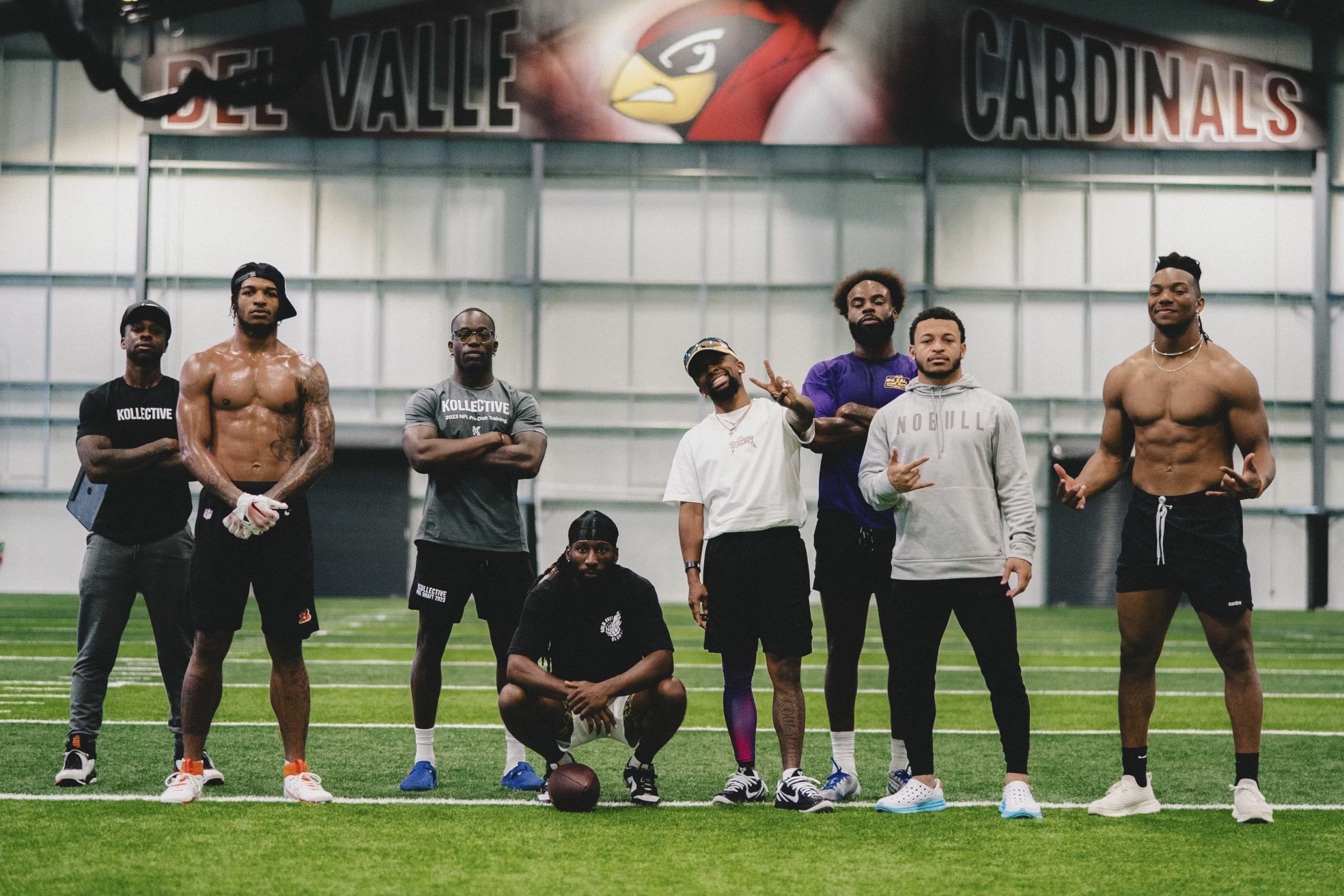 Group of seven men standing and kneeling inside an indoor sports facility, with a banner and sports equipment visible in the background.