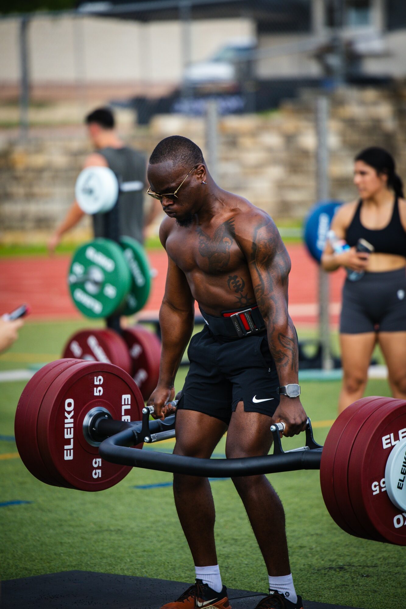 A shirtless man with glasses and tattoos lifts a heavy barbell outdoors on a sports field, with people in the background.