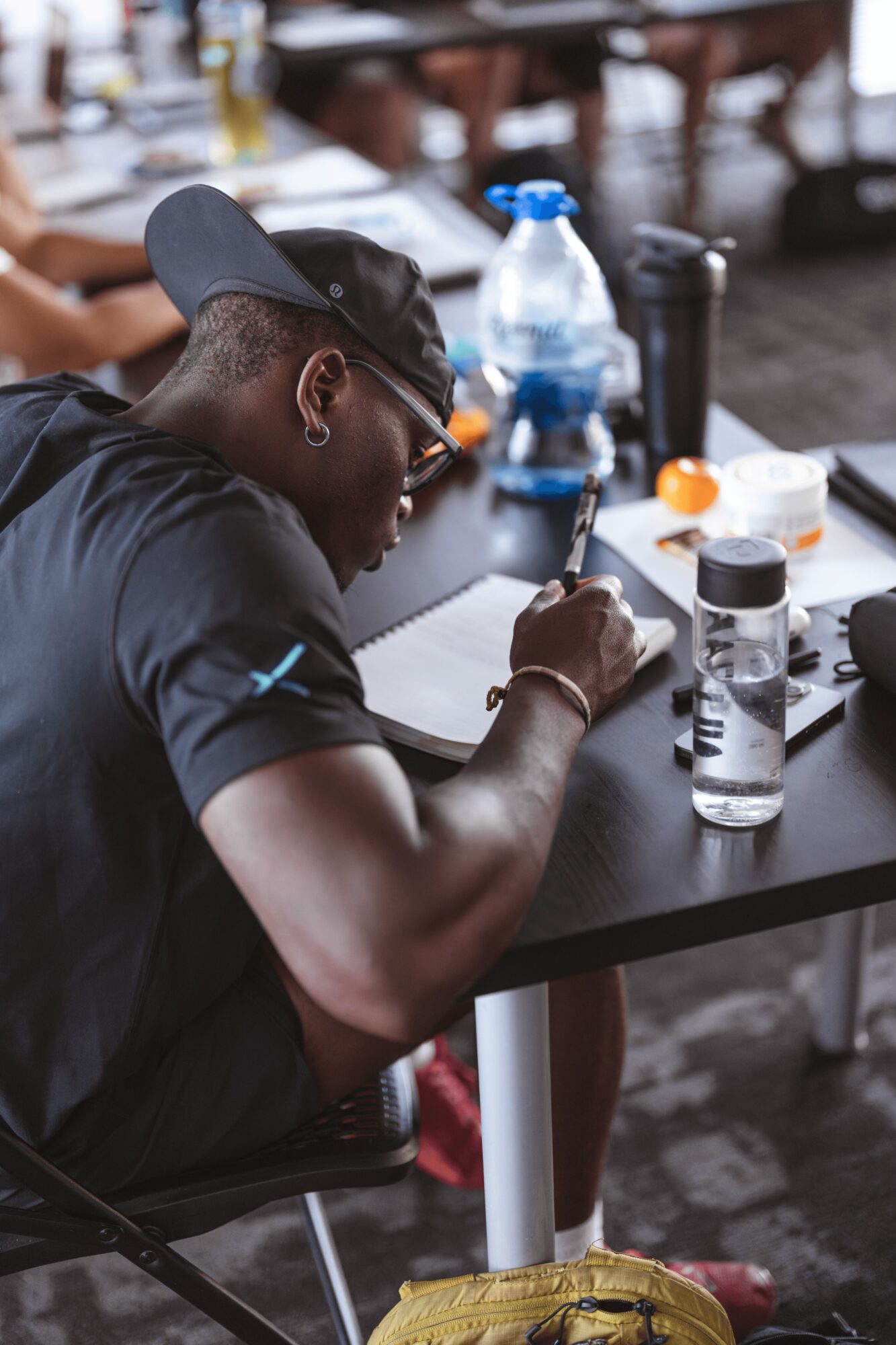 Person wearing a black cap and shirt writing on paper at a table with bottles and containers, in an outdoor setting.