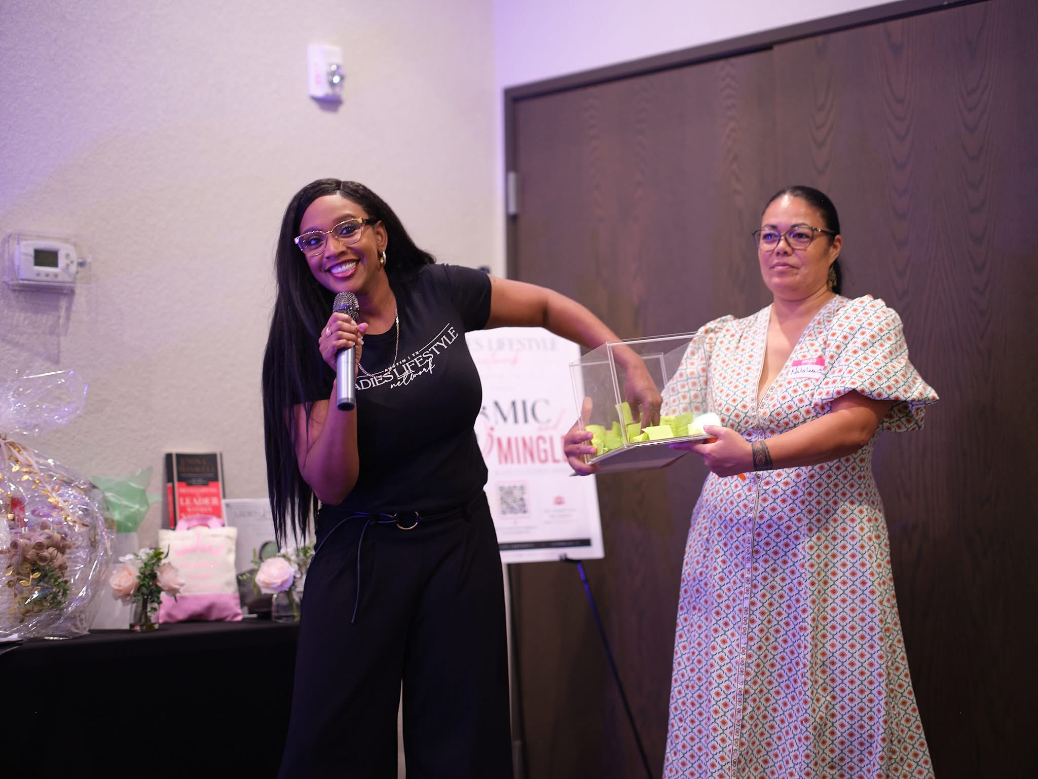 Two women standing indoors, one speaking into a microphone, the other holding a tray with a cake, smiling.
