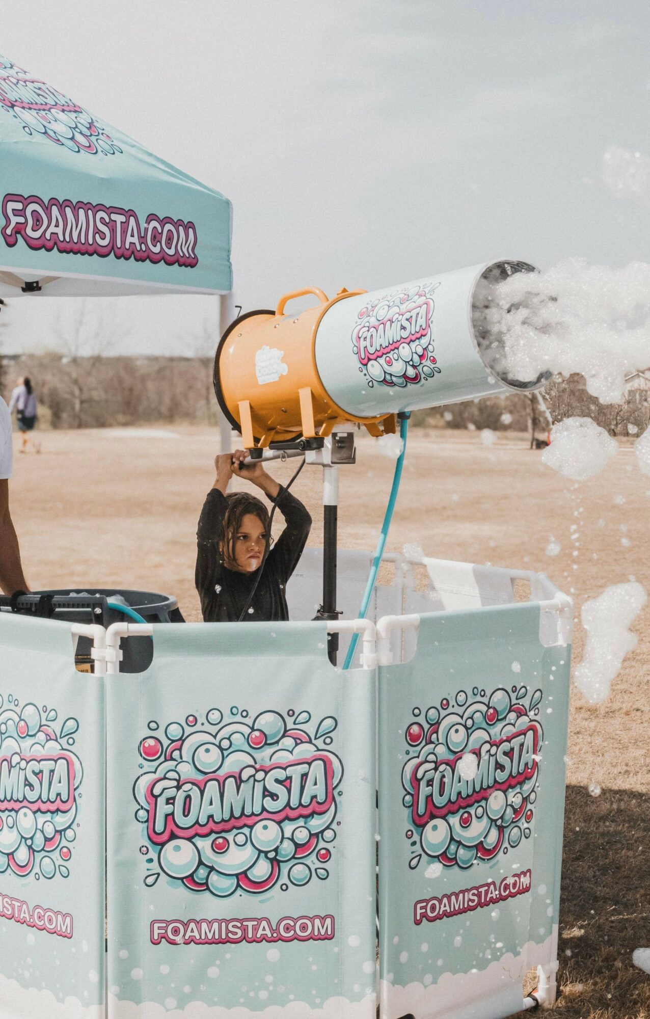 Child operating a foam cannon at foam party, with foam spraying out, surrounded by foam barriers with foamista branding.
