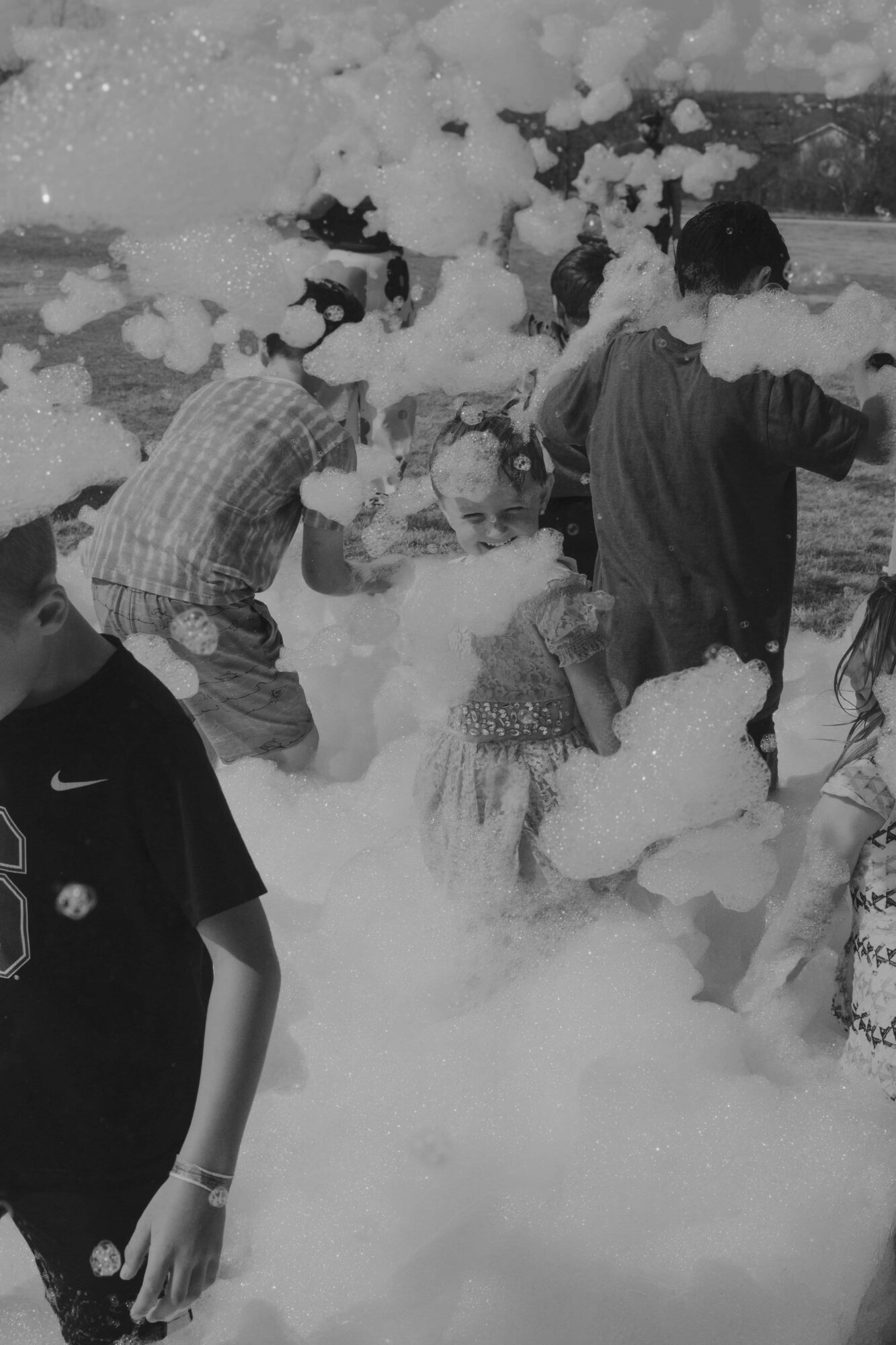 People playing in foam or bubbles outdoors, some with arms raised, surrounded by foam and bubbles.
