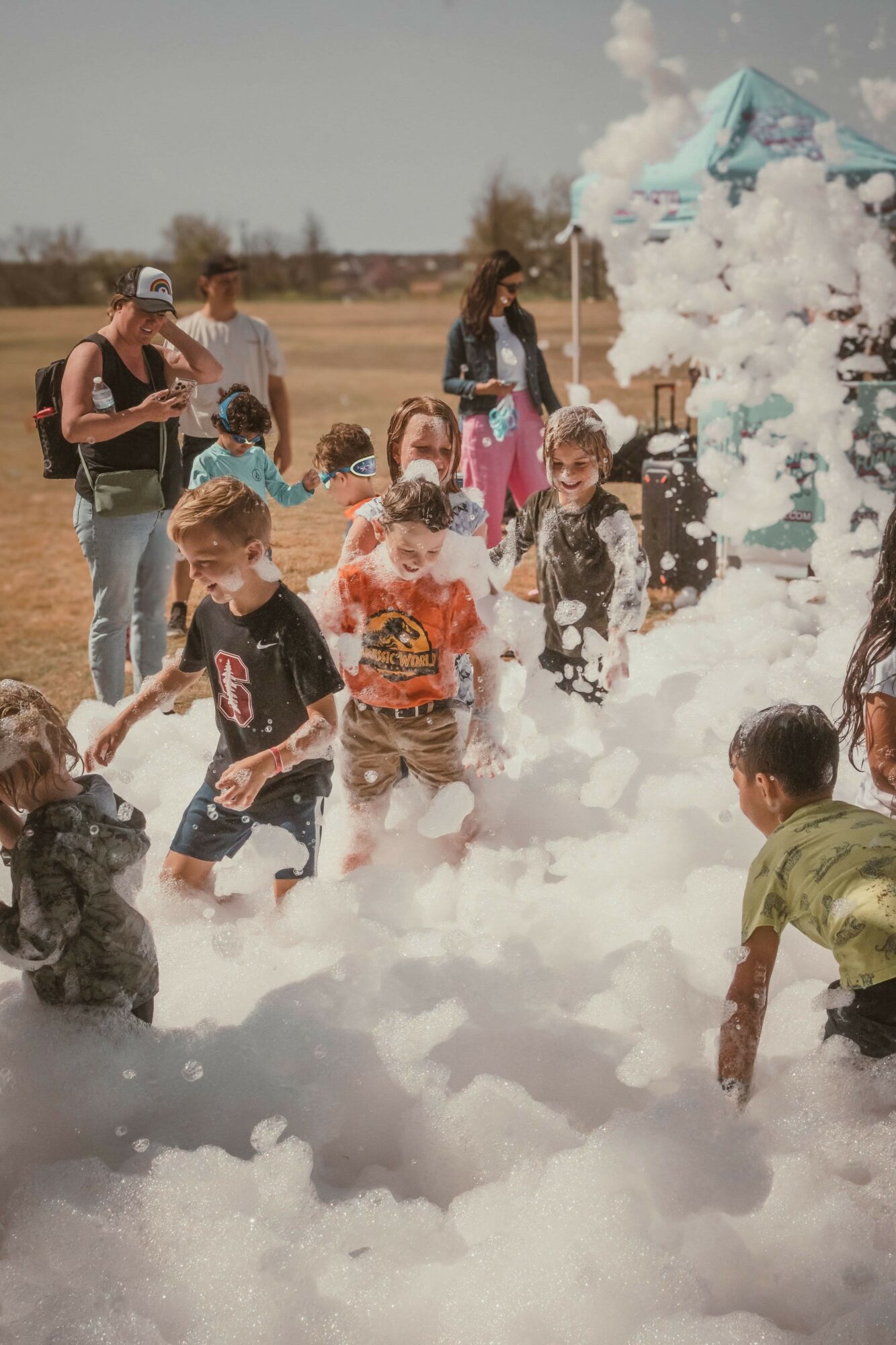 Children playing in foam outdoors with adults watching, some children are smiling and running through foam, open field background.