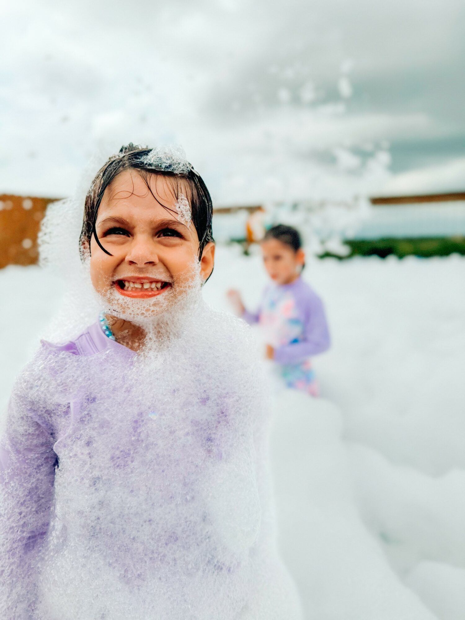 Child smiling with foam on face, other child in background, outdoors with cloudy sky.