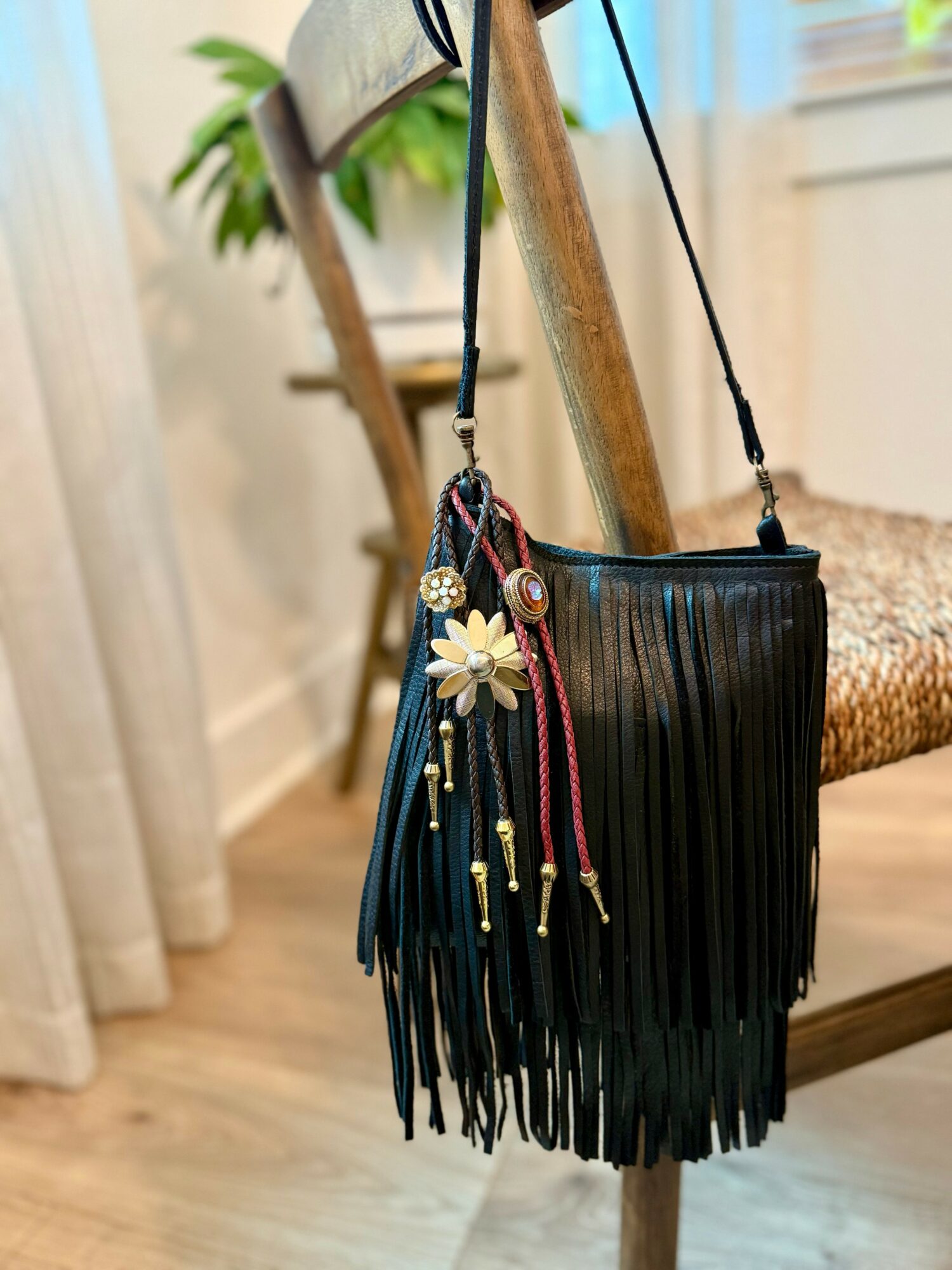 Black fringed handbag hanging from a wooden chair with a decorative flower and beads, indoors near a window.