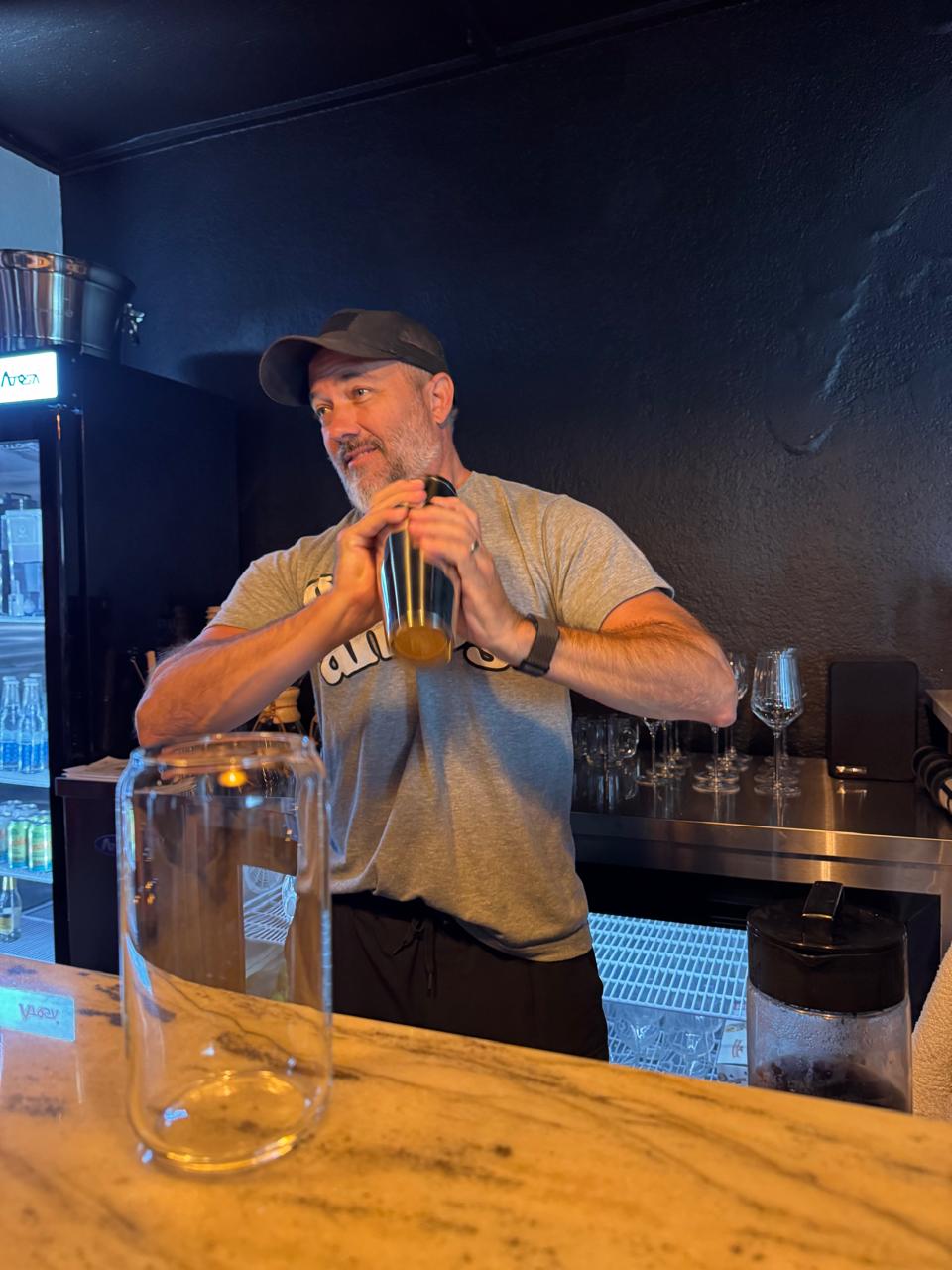Man wearing a cap and gray T-shirt holding a bottle, standing behind a bar counter with a glass jar in front.