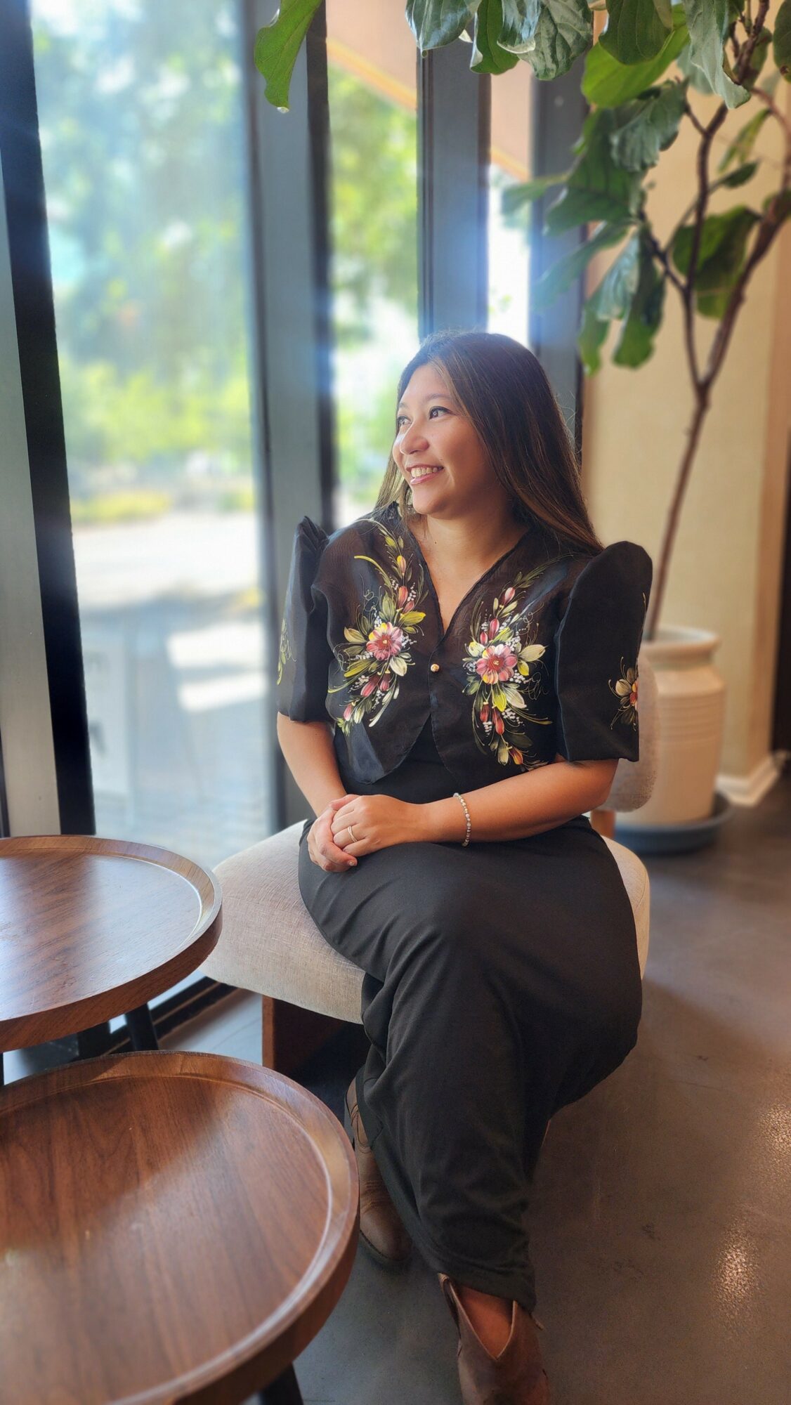 Woman sitting by window with greenery outside, smiling and looking to the side, wearing a black embroidered top and gray pants.