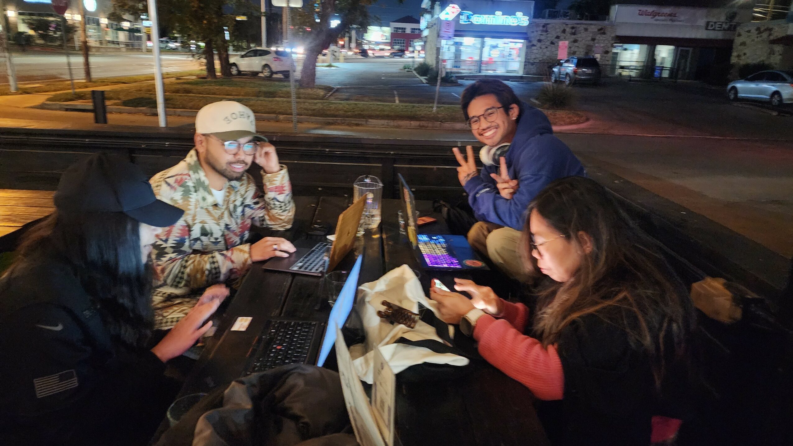 Four people sitting at a table outdoors at night, using laptops and smartphones, with a city street in the background.