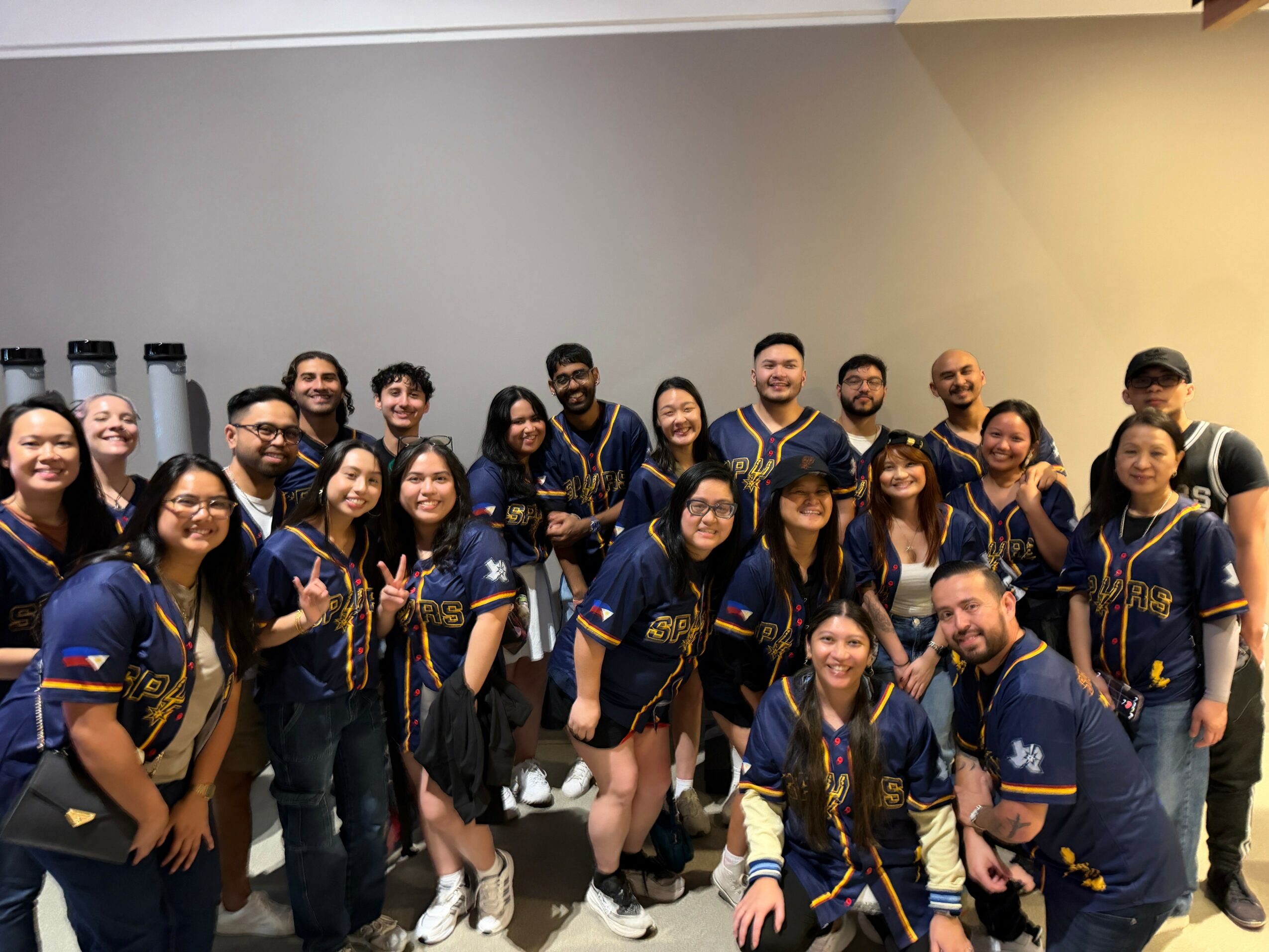 Group of people wearing matching navy blue and gold jackets, standing indoors against a plain wall, smiling and posing for the photo.
