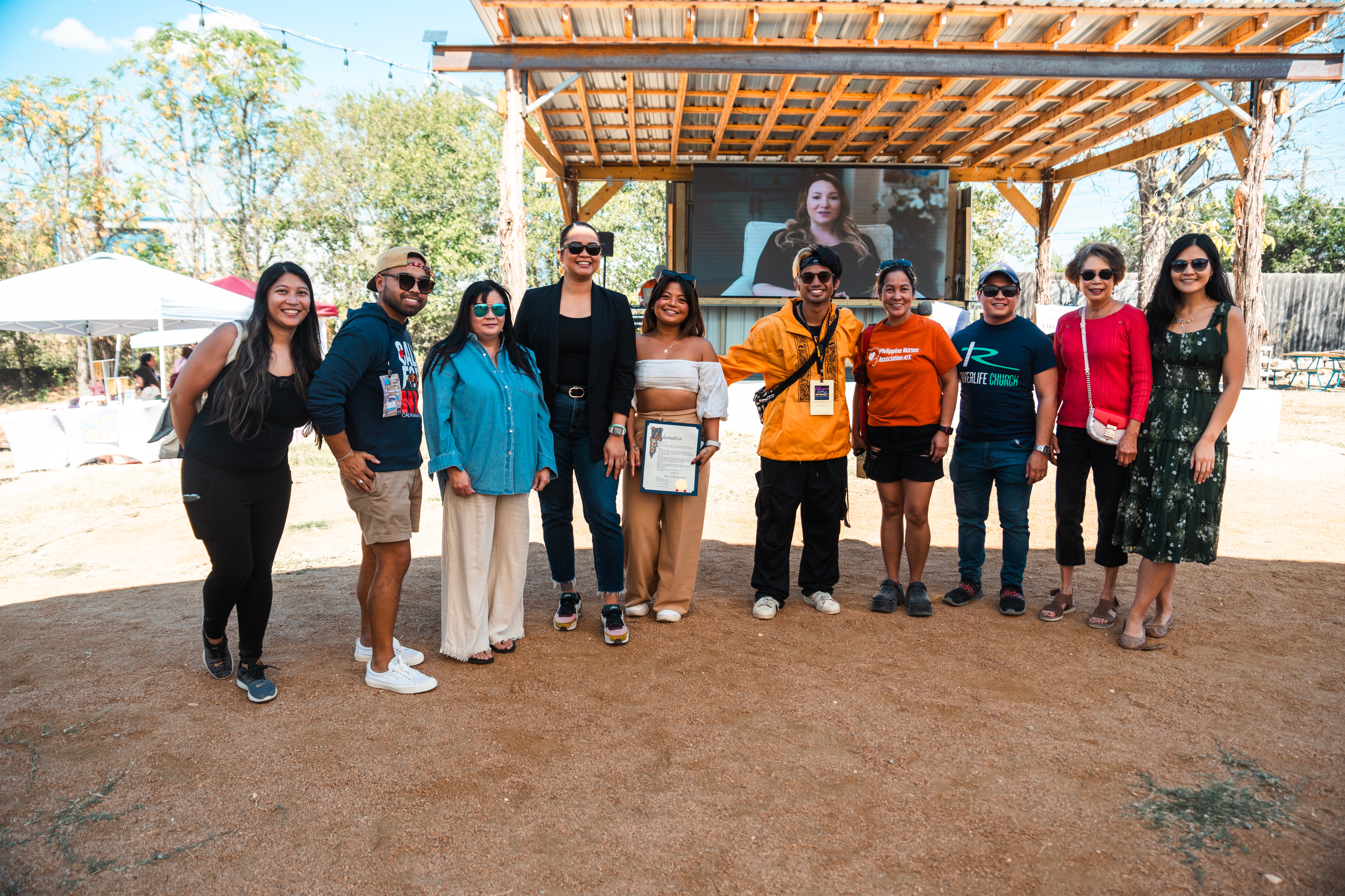 Group of ten people standing outdoors in front of a stage with a large screen, trees, and tents in the background.