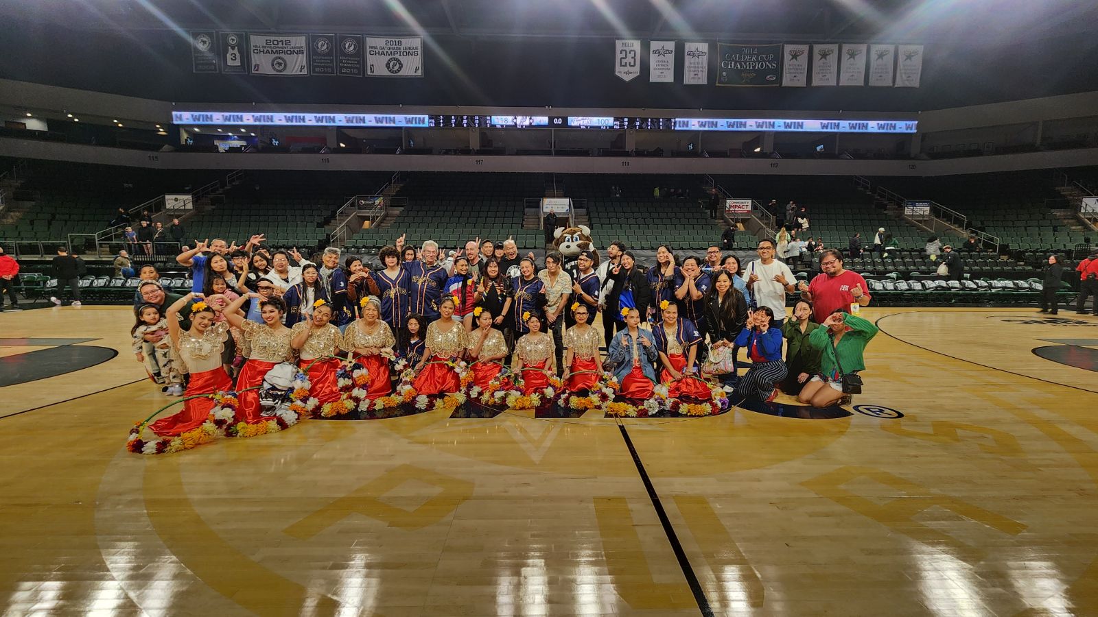 Group of people, including children in costumes, gathered on a basketball court indoors, some sitting and some standing.