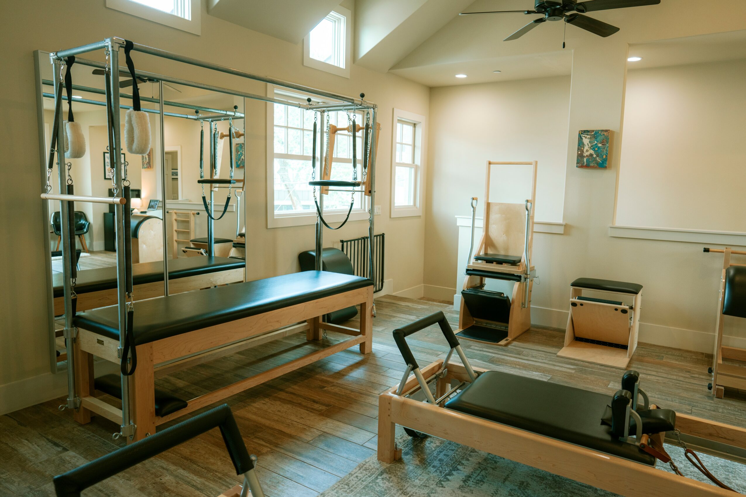 Rehabilitation or physical therapy room with equipment, windows, and wooden flooring, featuring a Pilates reformer and other exercise machines.