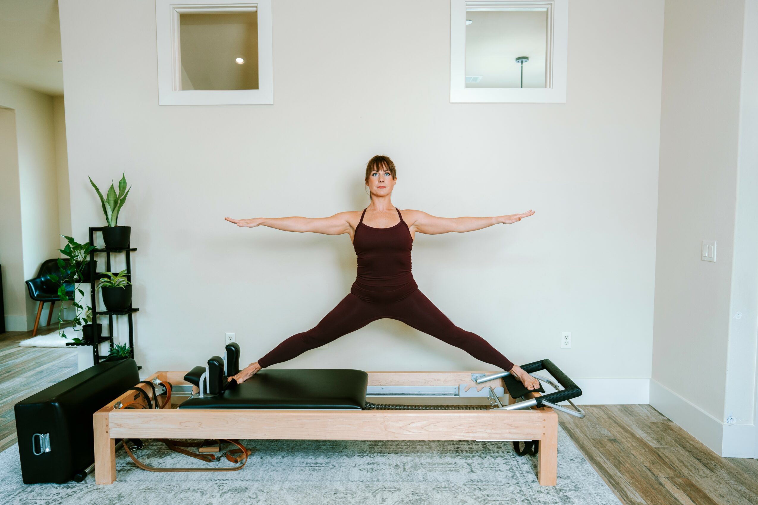 Woman in a yoga pose with arms and legs extended, standing on a wooden frame in a bright room.