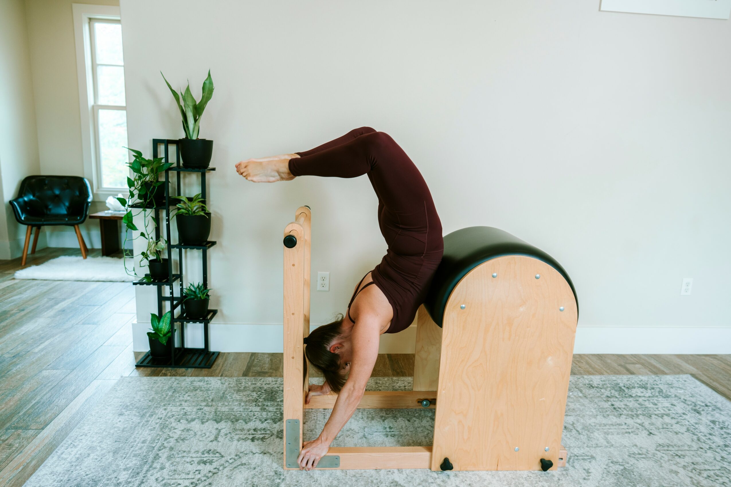 Person performing a backbend on a wooden yoga prop in a bright room with plants and furniture.
