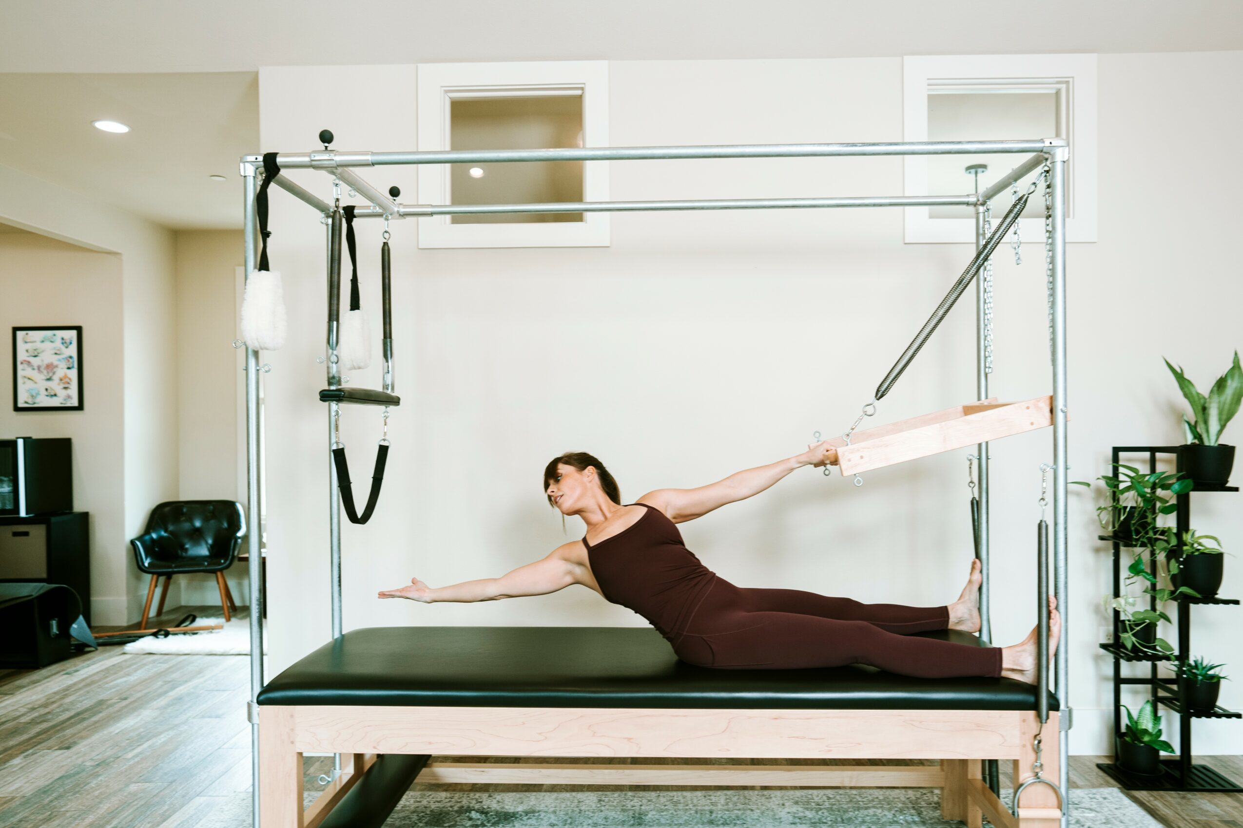 Woman performing a stretch on a specialized exercise table in a bright room with plants and furniture.