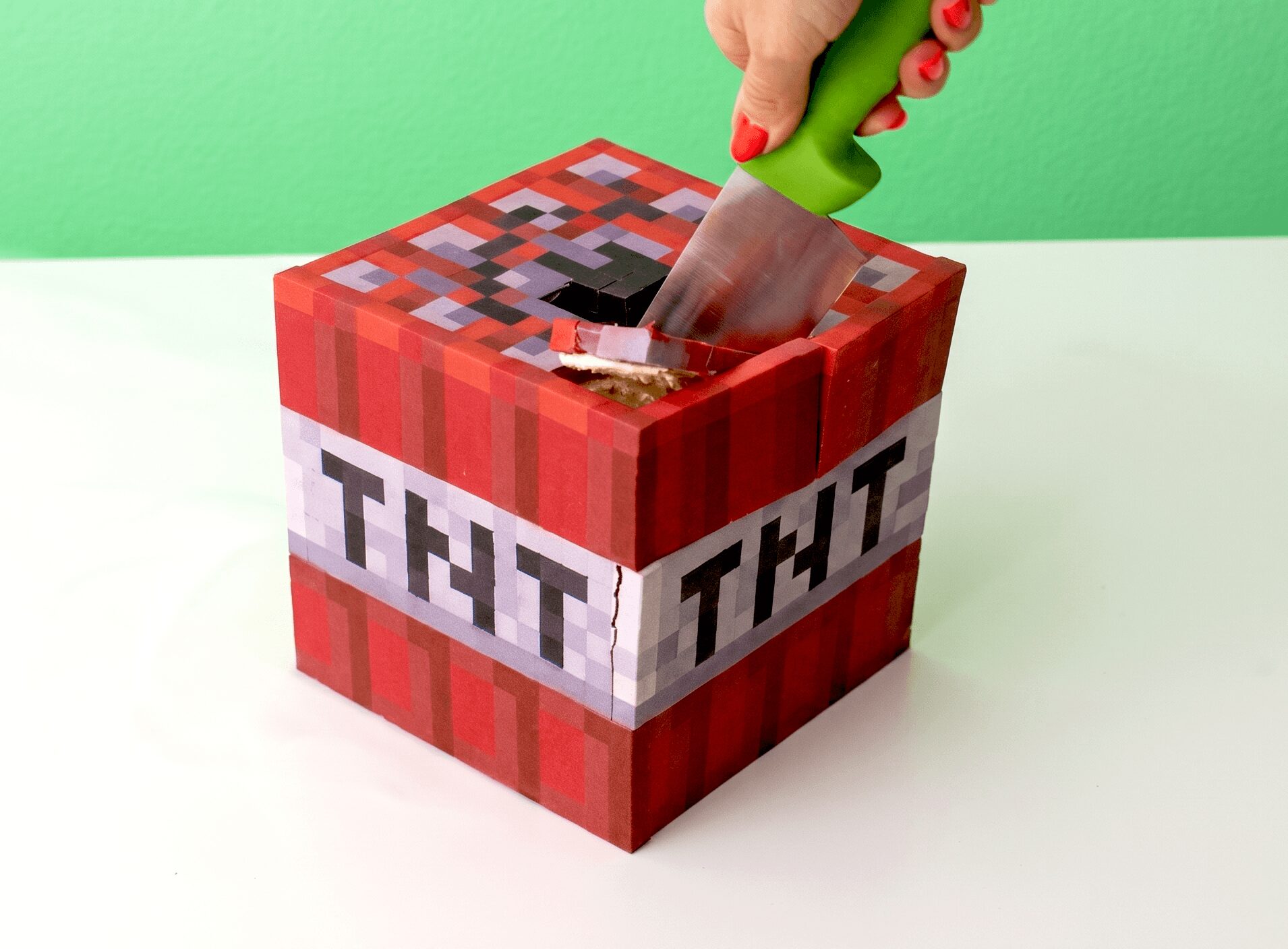 Hand placing a knife into a Minecraft-themed TNT box with a green background, white surface, and pixelated design.