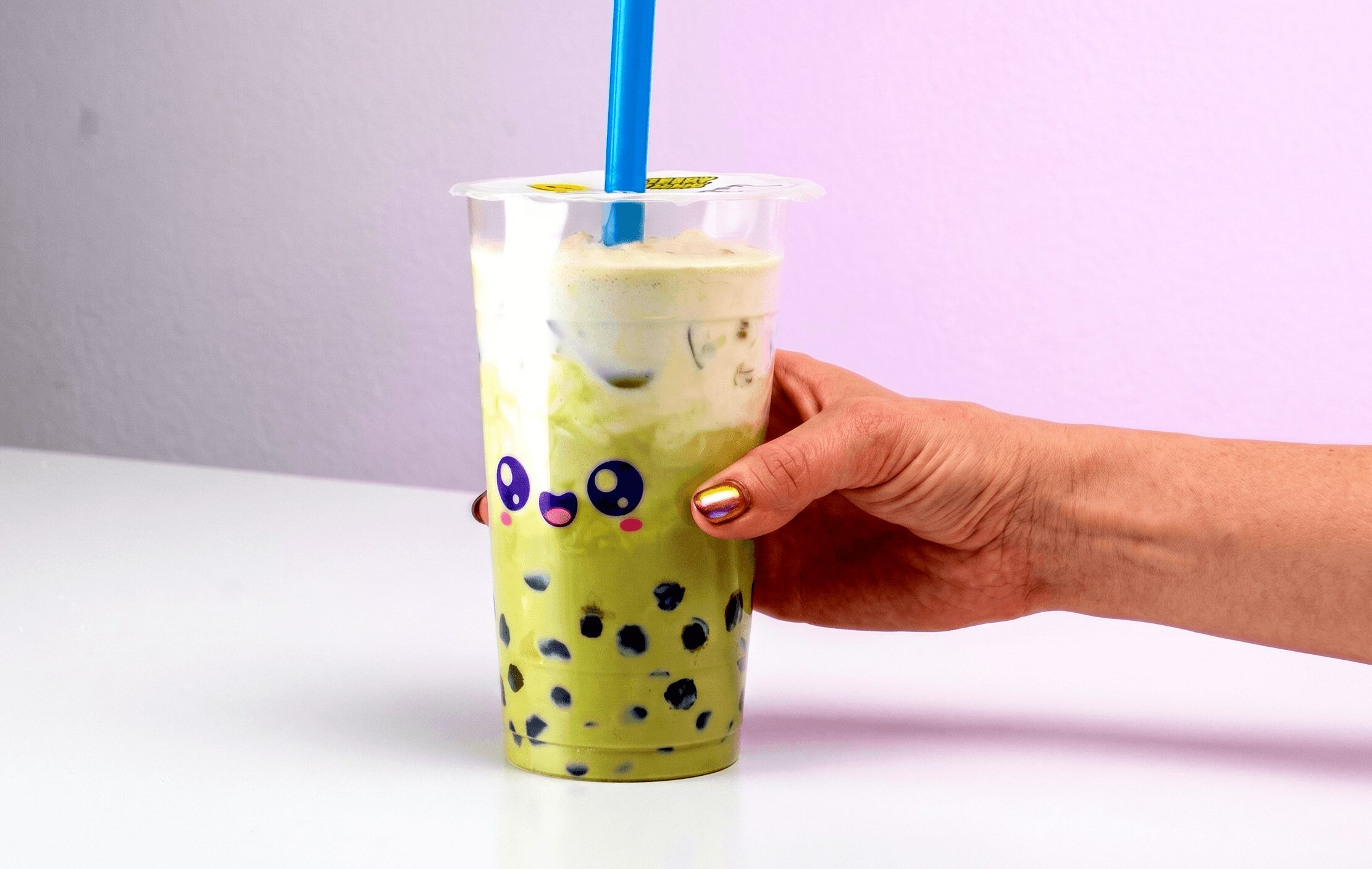 Hand holding a plastic cup with a yellow-green beverage and black tapioca pearls, with a blue straw, against a plain background.