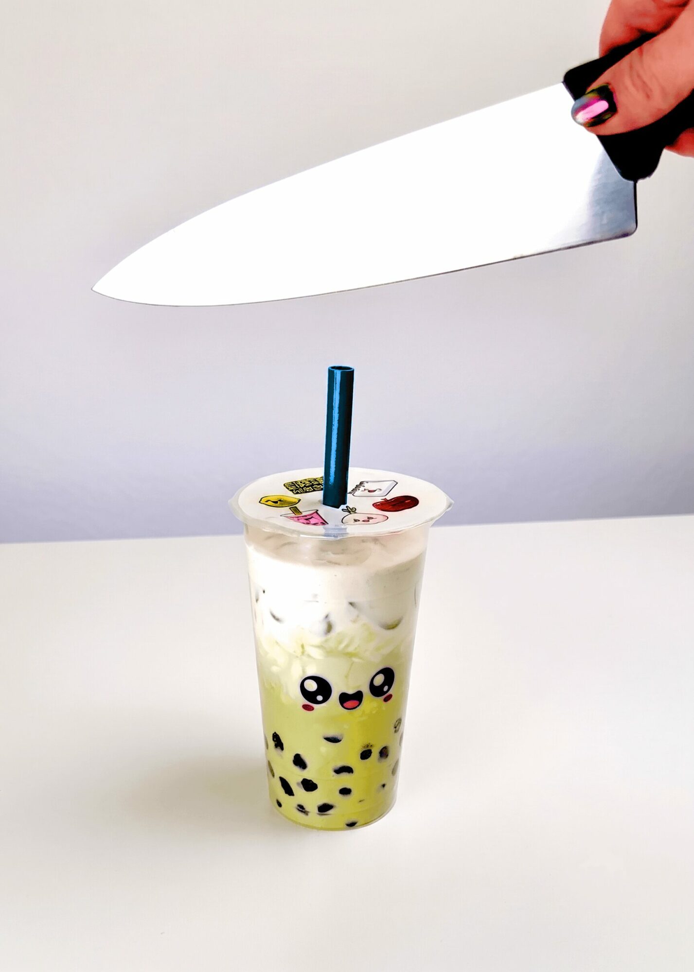 A hand holds a large knife above a bubble tea with a cartoon face, on a white surface and background.