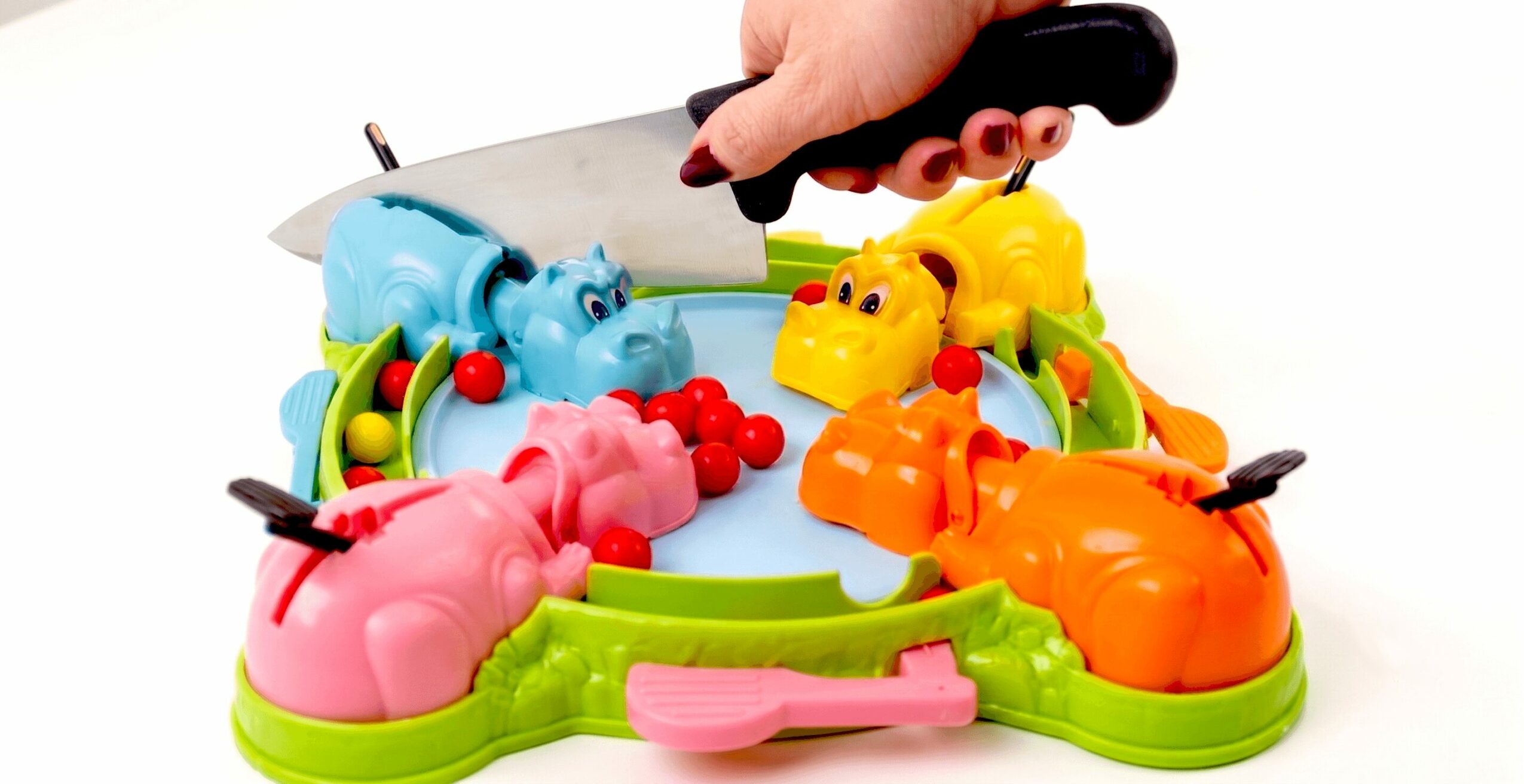 Child's hand holding a knife over a colorful plastic crocodile game with animal figures and red balls.