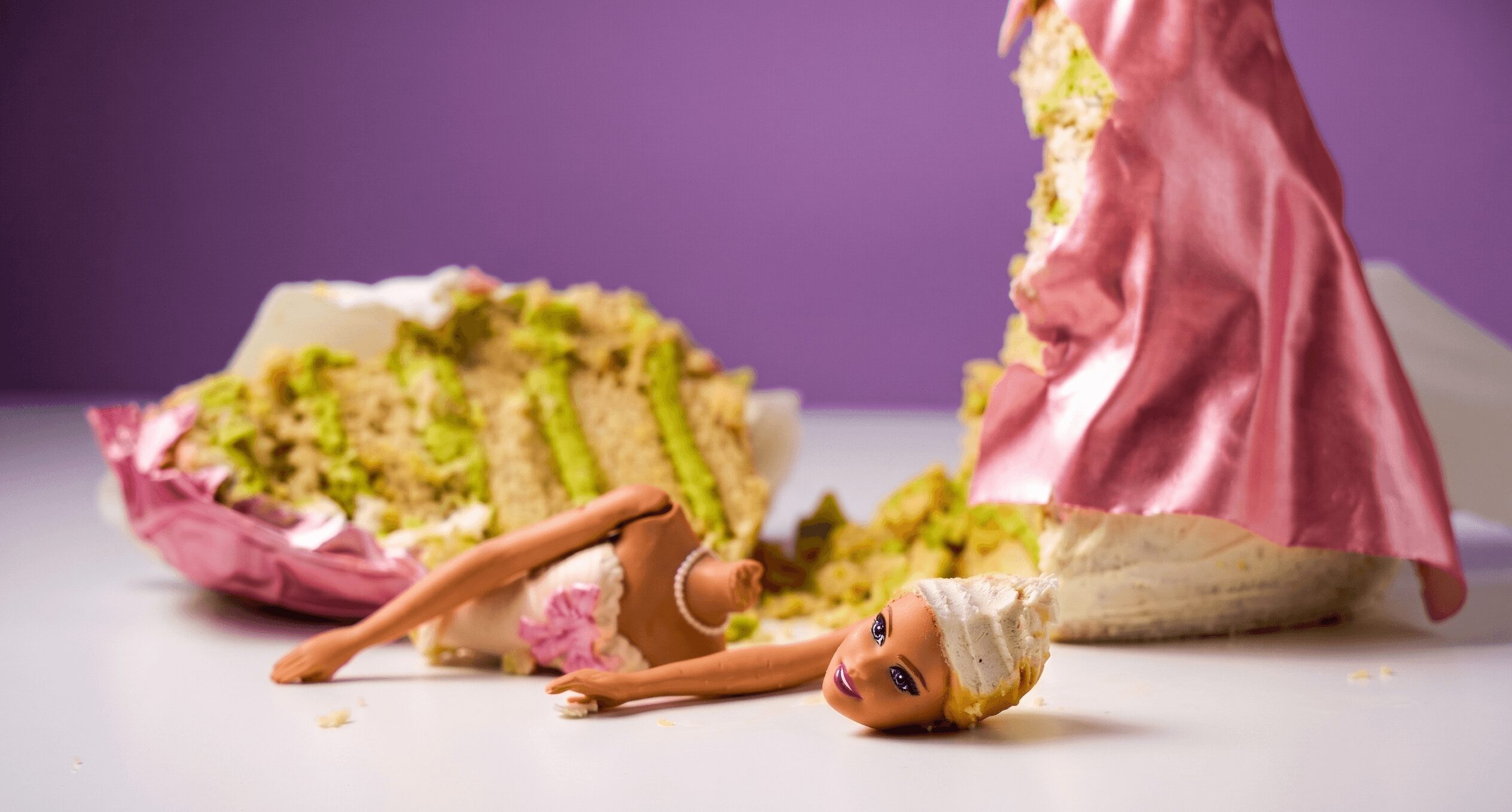 Doll lying on its side with a broken cake in the background, on a white surface with a purple backdrop.