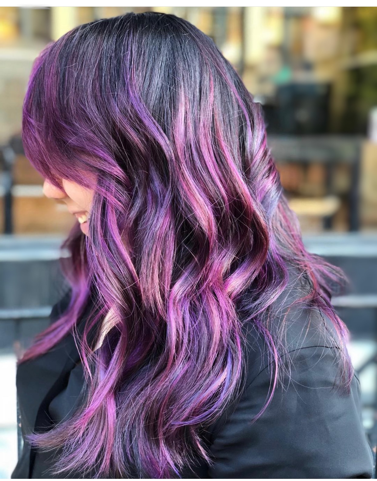 Side view of a woman with wavy hair dyed in shades of purple and pink, outdoors with blurred background.