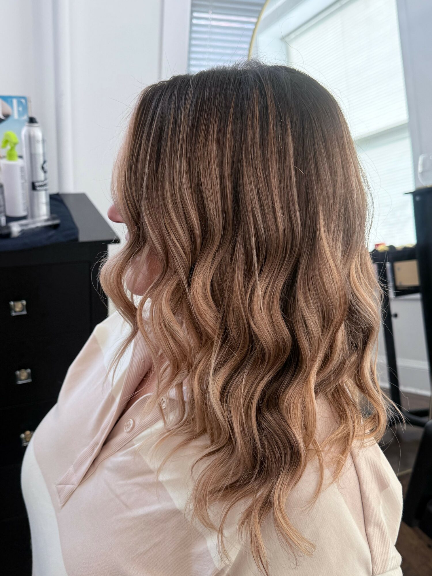 Side view of a woman with long, wavy, light brown hair, wearing a light-colored top, indoors with a window in the background.