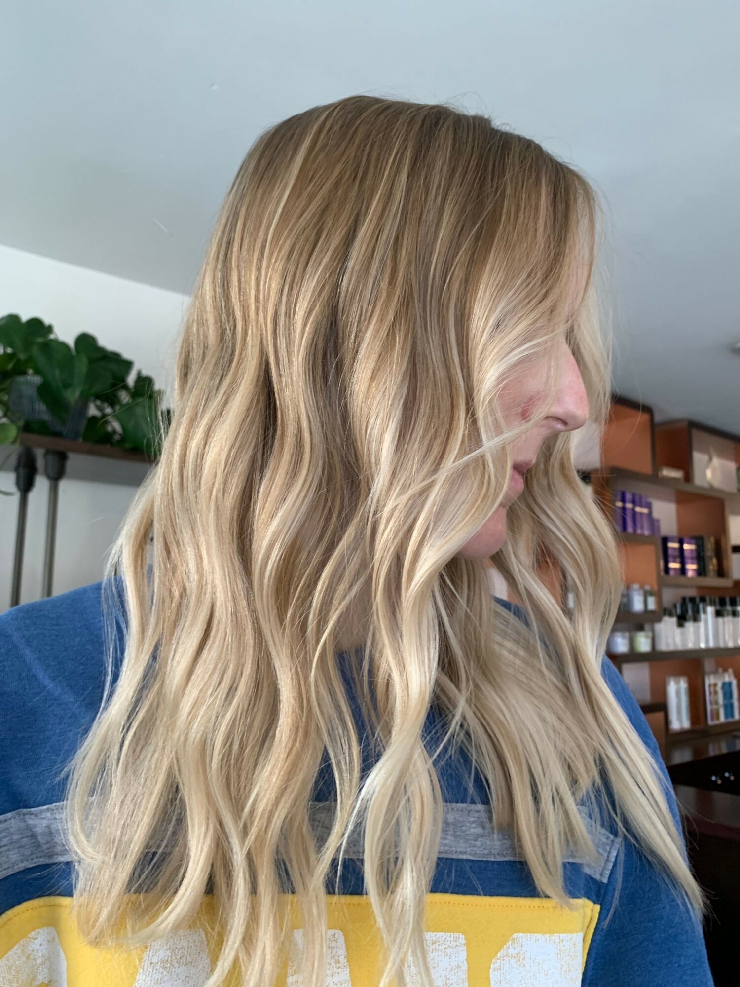 Side view of a person with long, wavy blonde hair wearing a blue and yellow shirt, indoors with shelves in background.