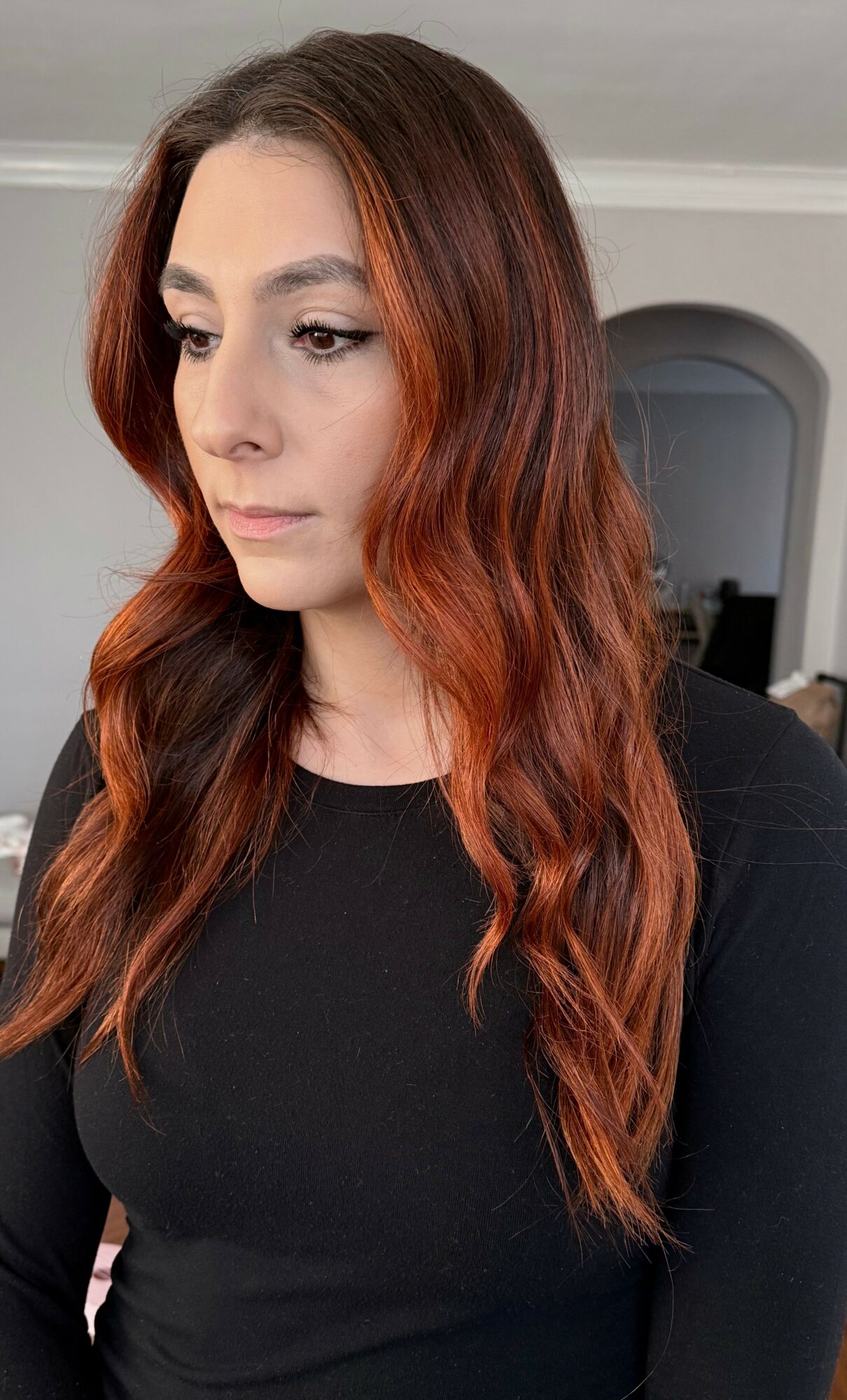 Woman with long, wavy reddish-brown hair wearing a black top, looking to the side indoors.