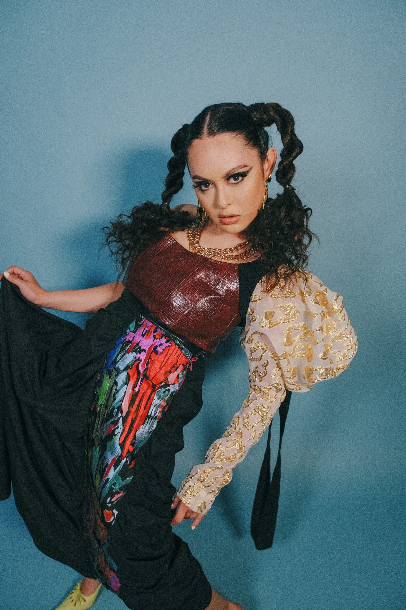 Woman with styled hair and makeup, wearing a shiny top, lace sleeve, and colorful skirt, posing against a blue background.