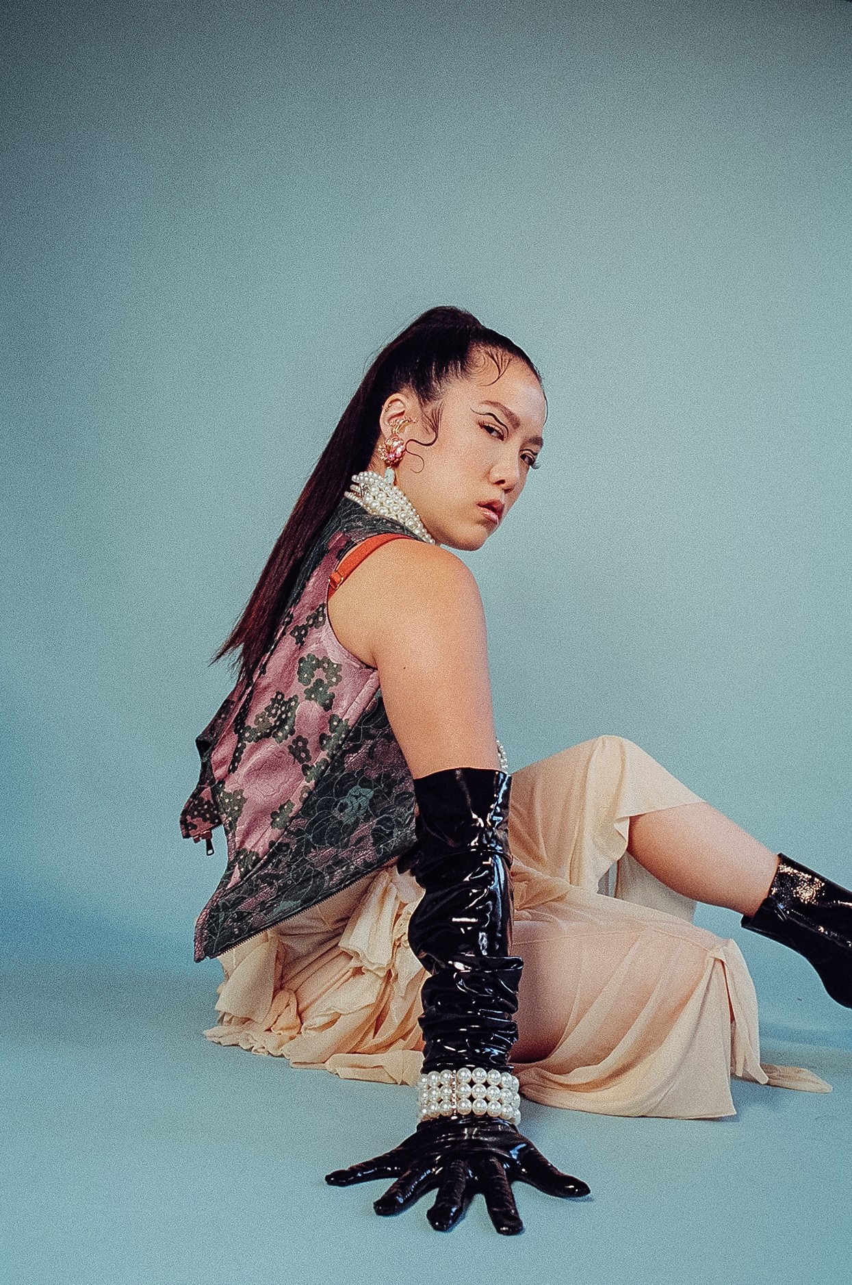 Young woman with long dark hair sitting on the floor against a blue background, wearing a patterned top, beige pants, and black gloves.