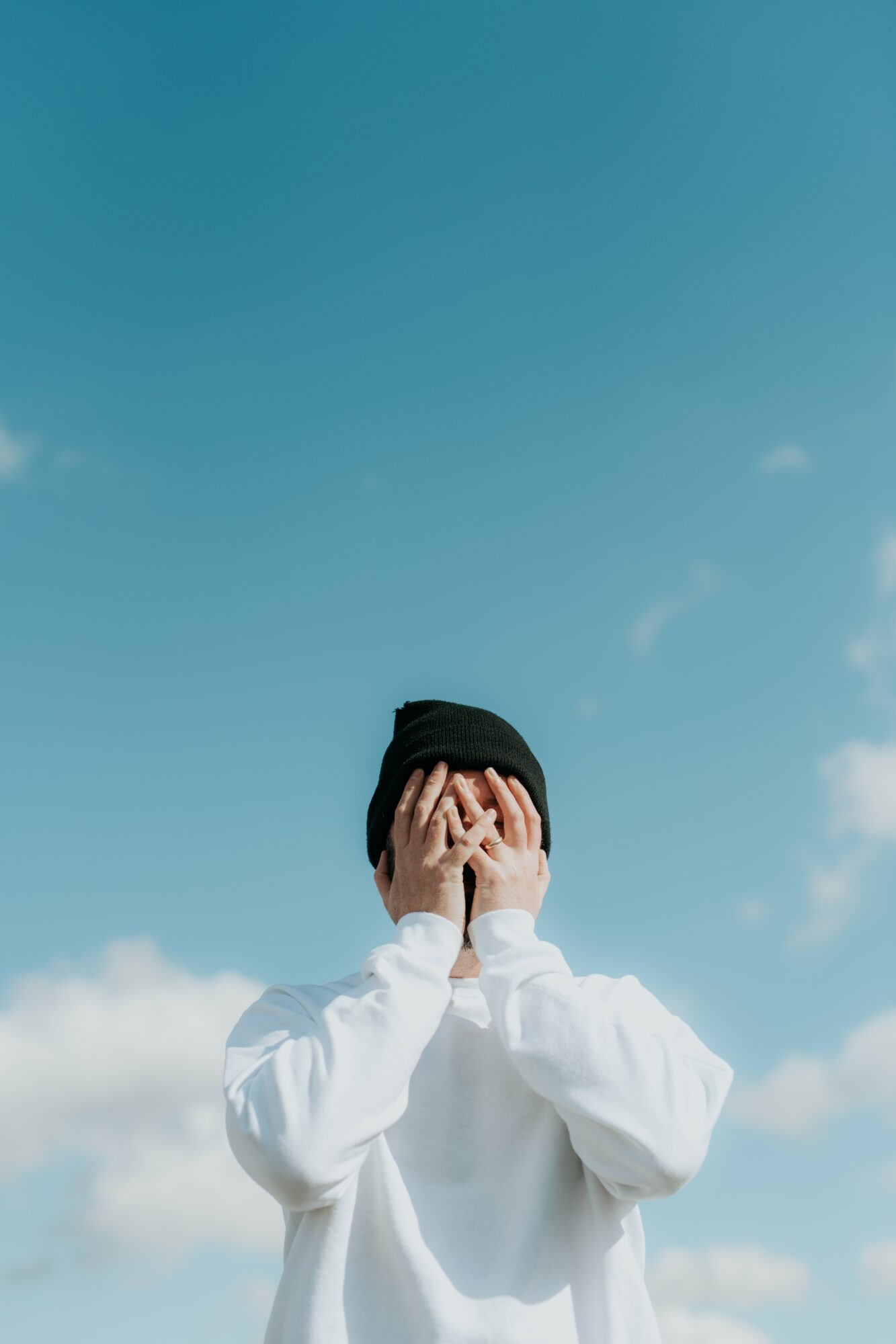 Person wearing a white hoodie and black beanie covers their face with hands against a blue sky with clouds